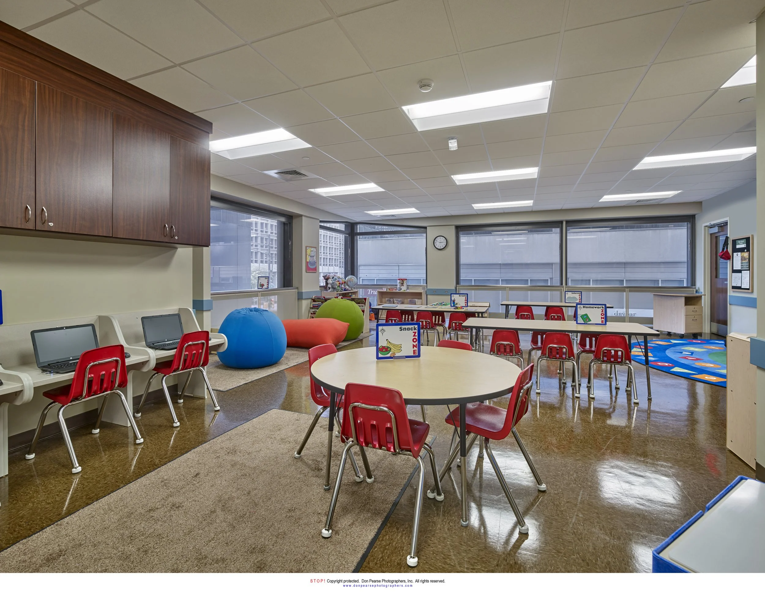 Empty classroom with tables and chairs, laptops on side tables, large windows, colorful bean bags, and a blue rug with a play area.