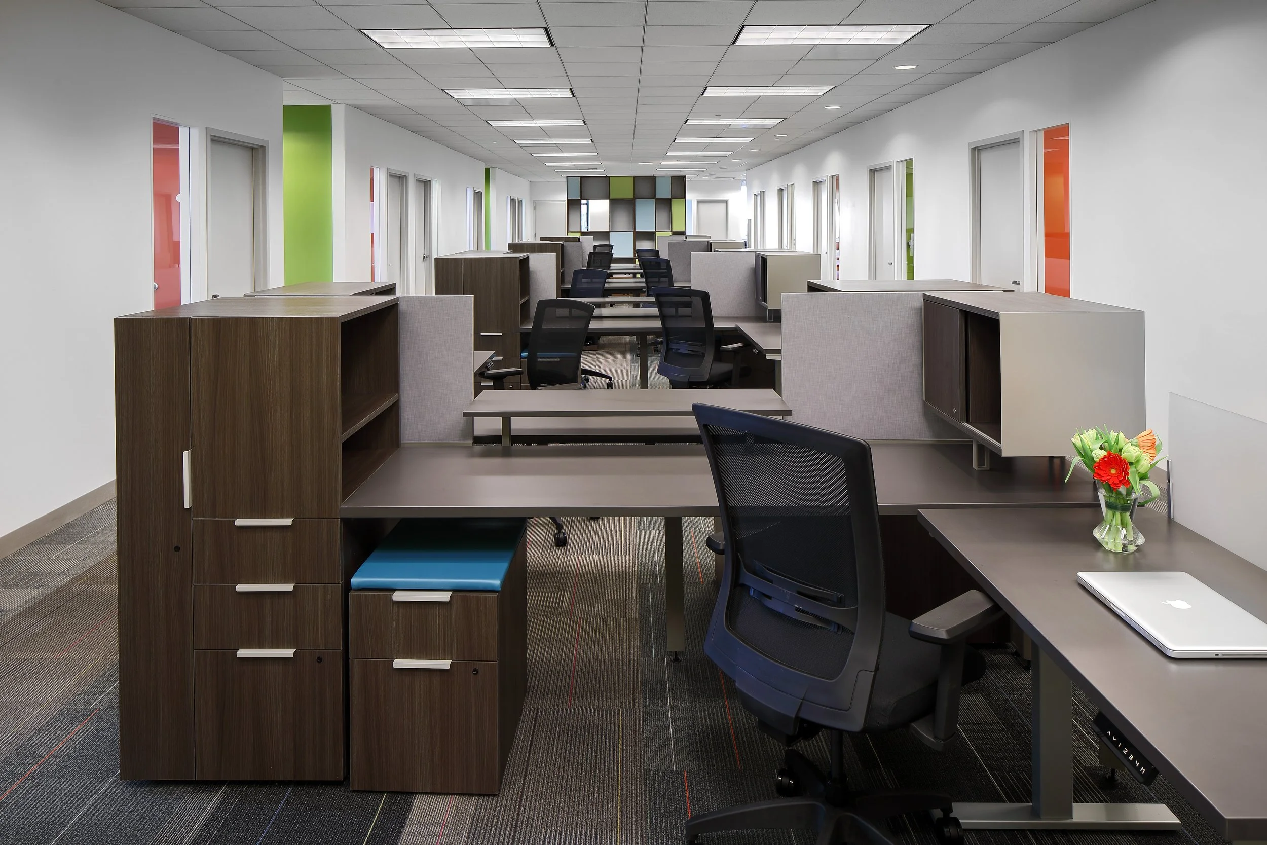 Empty modern office workspace with multiple desks, chairs, and colorful glass doors along the corridor.