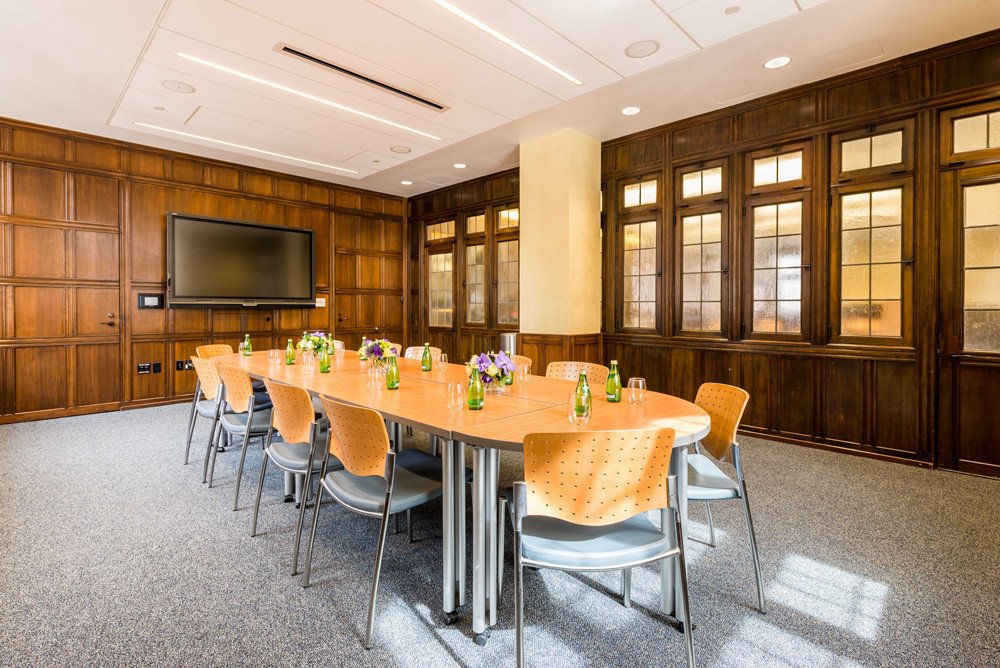 A conference room with a long oval table, orange chairs, and a large flat-screen TV on a wood-paneled wall. The table has bottles of water and floral arrangements, with wooden framed windows along one wall.