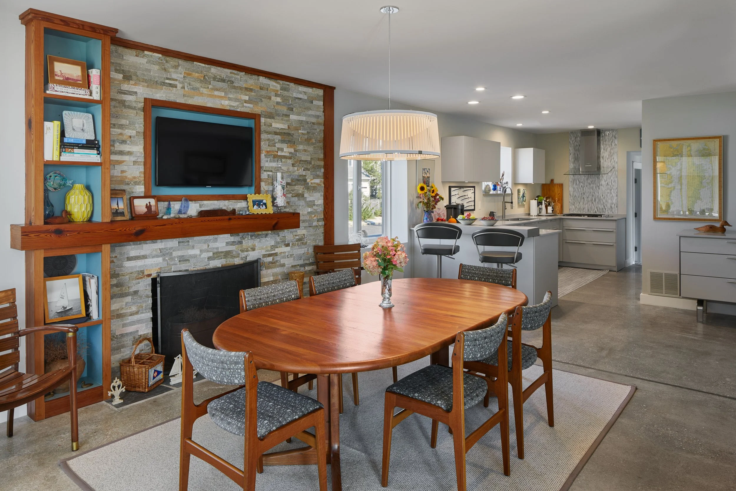 Open-concept dining area and kitchen with stone fireplace, flat-screen TV, and various decorative items, including flowers and framed pictures.