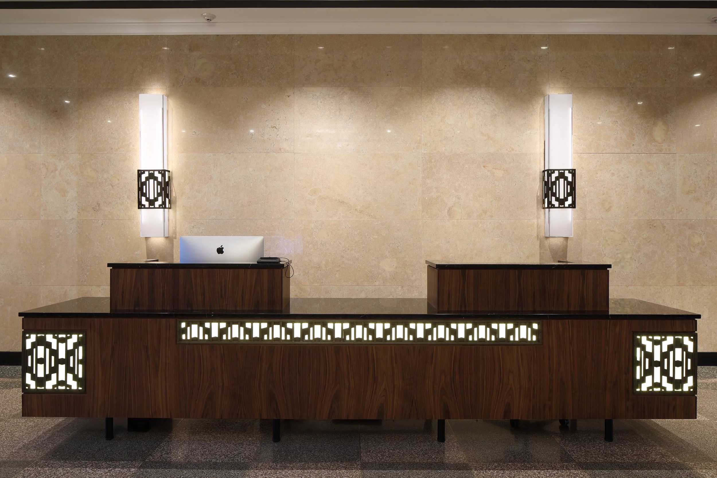 Empty hotel reception desk with a computer, illuminated decorative panels, and wall-mounted lights against a beige marble wall.