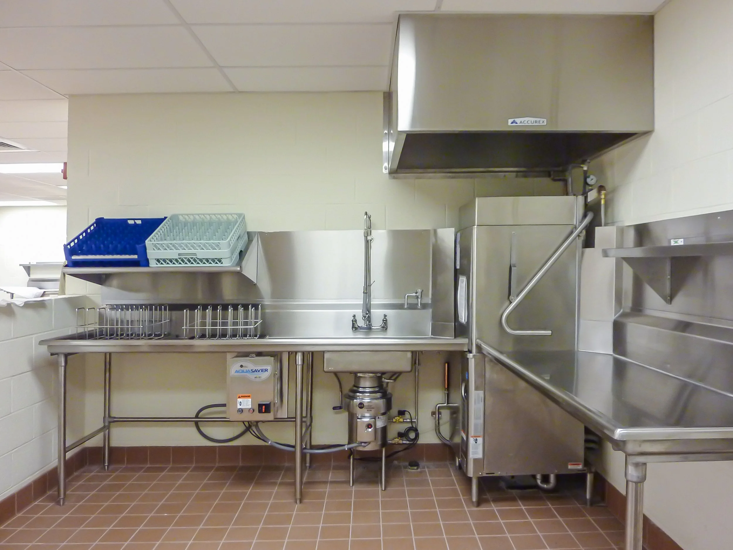 Stainless steel commercial kitchen sink with dishes rack and industrial dishwasher in a food service area.