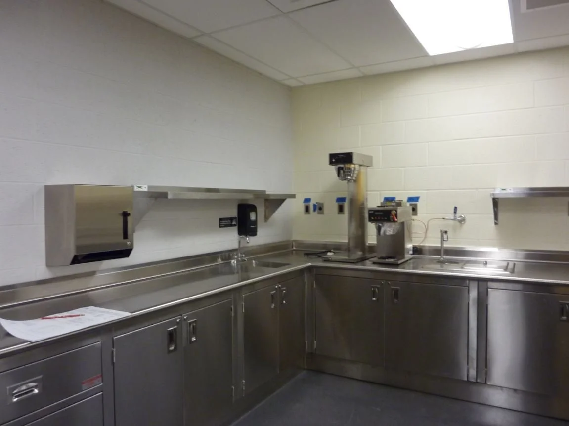 Empty stainless steel commercial kitchen sink with shelves, a paper towel dispenser, and a coffee machine in the corner.