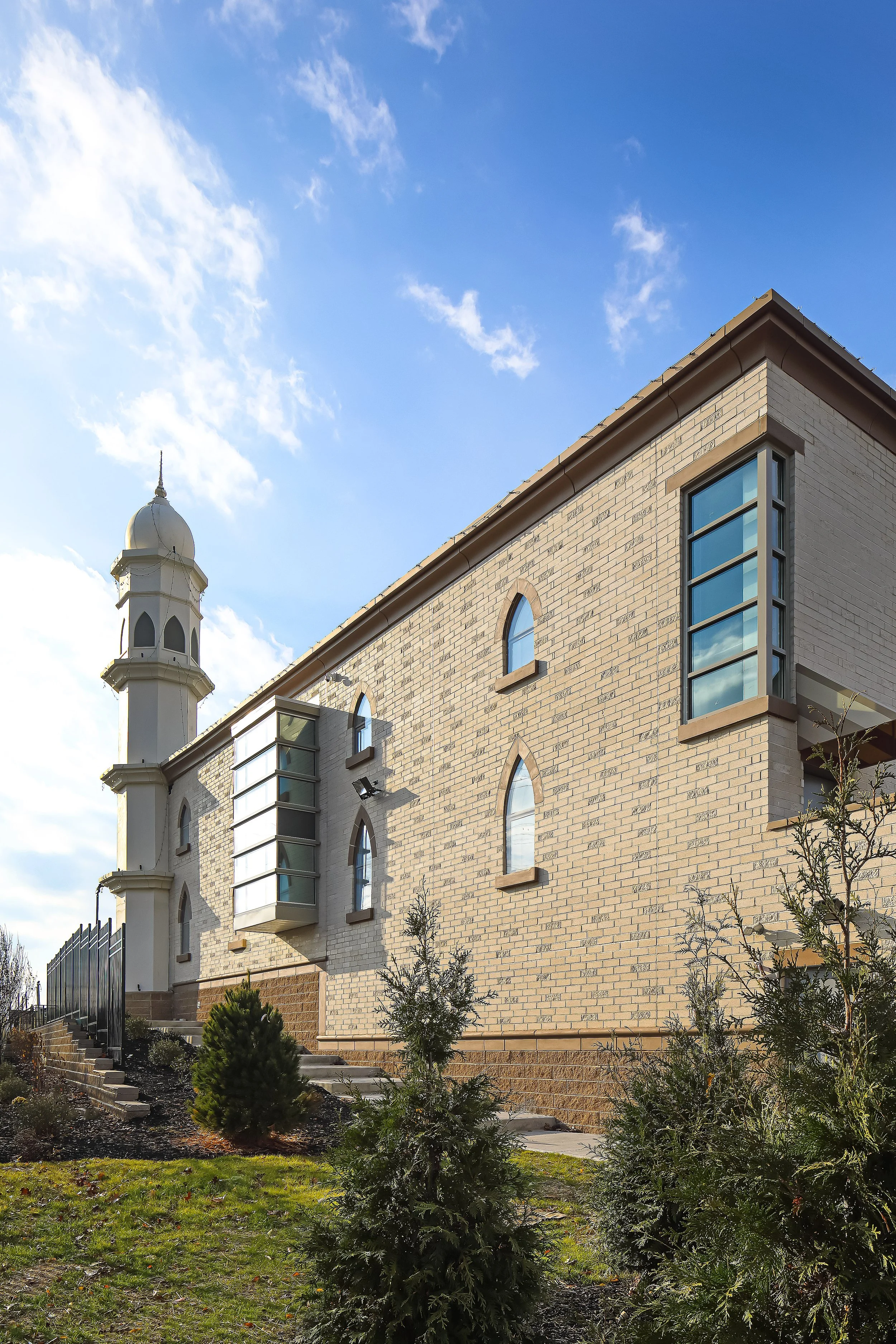 A beige brick building with arched windows and modern glass extensions, topped with a small turret, under a blue sky with clouds, surrounded by small bushes and trees.