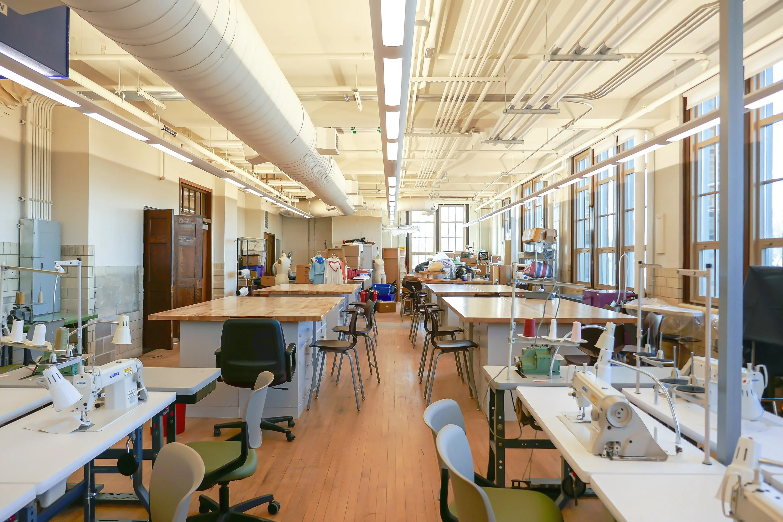 A spacious, well-lit workshop or studio with workbenches, sewing machines, mannequins, and chairs, illuminated by large windows on the right side and overhead lighting. The space has an industrial ceiling with exposed ducts.