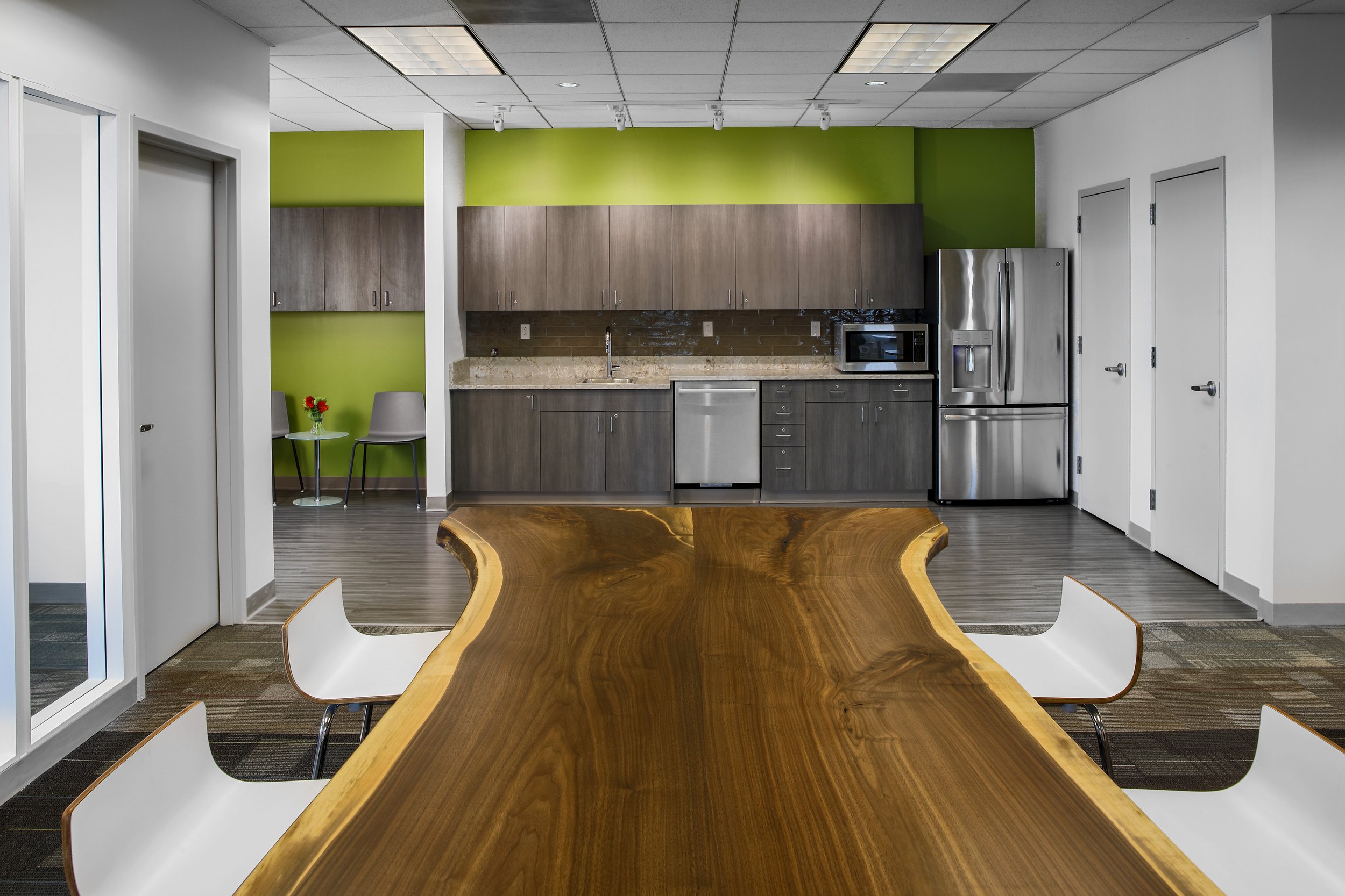 Modern kitchen with grey cabinets, stainless steel refrigerator, microwave, dishwasher, and a beige stone countertop; green accent wall; wooden table in the foreground and two chairs with white seats; small seating area with two chairs and a round ta