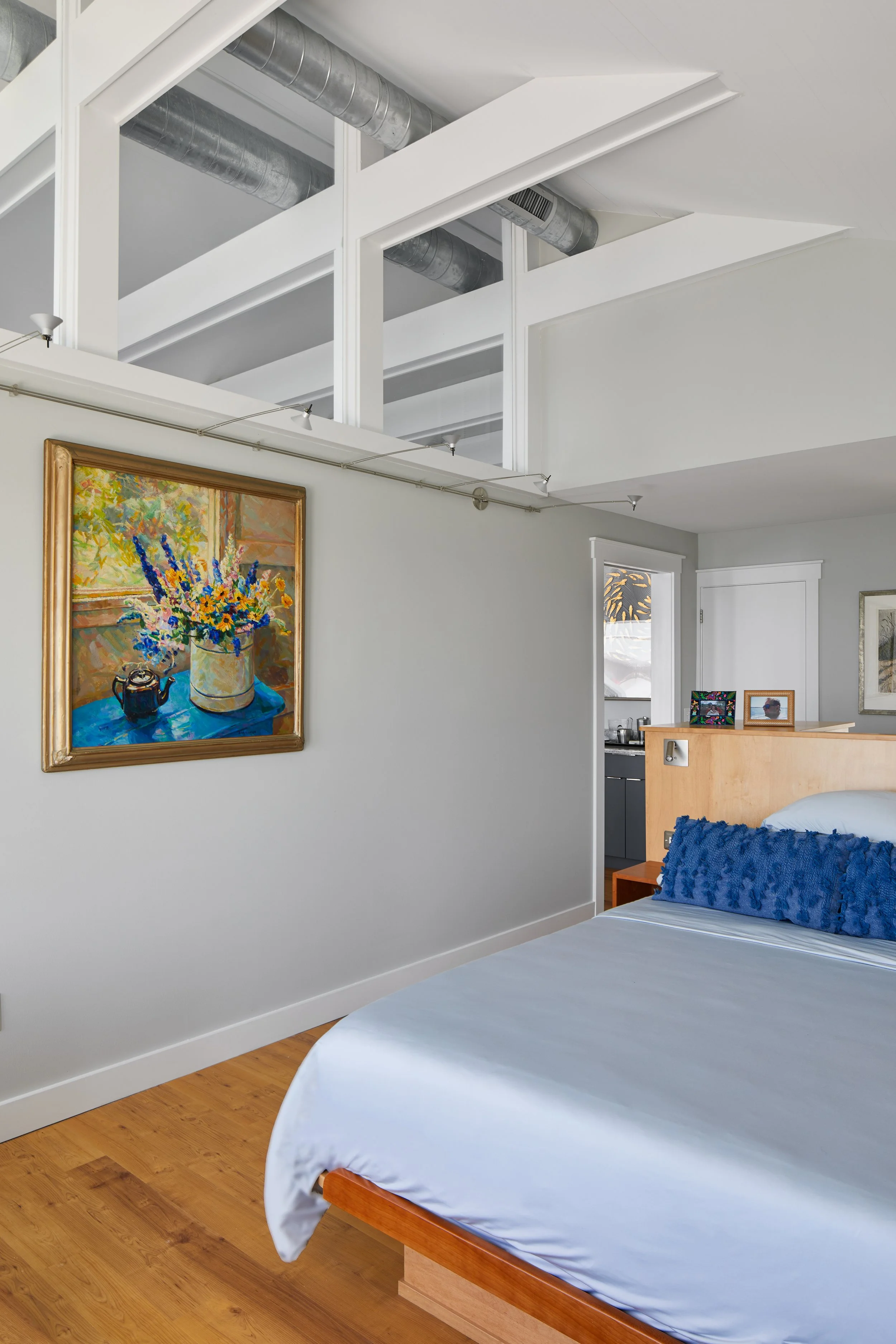 A bedroom with a bed that has a white sheet and a textured blue pillow. A wooden nightstand and a partial view of a room with a door and framed pictures. A wall with a colorful still life painting of a vase of flowers and a small kettle. The ceiling