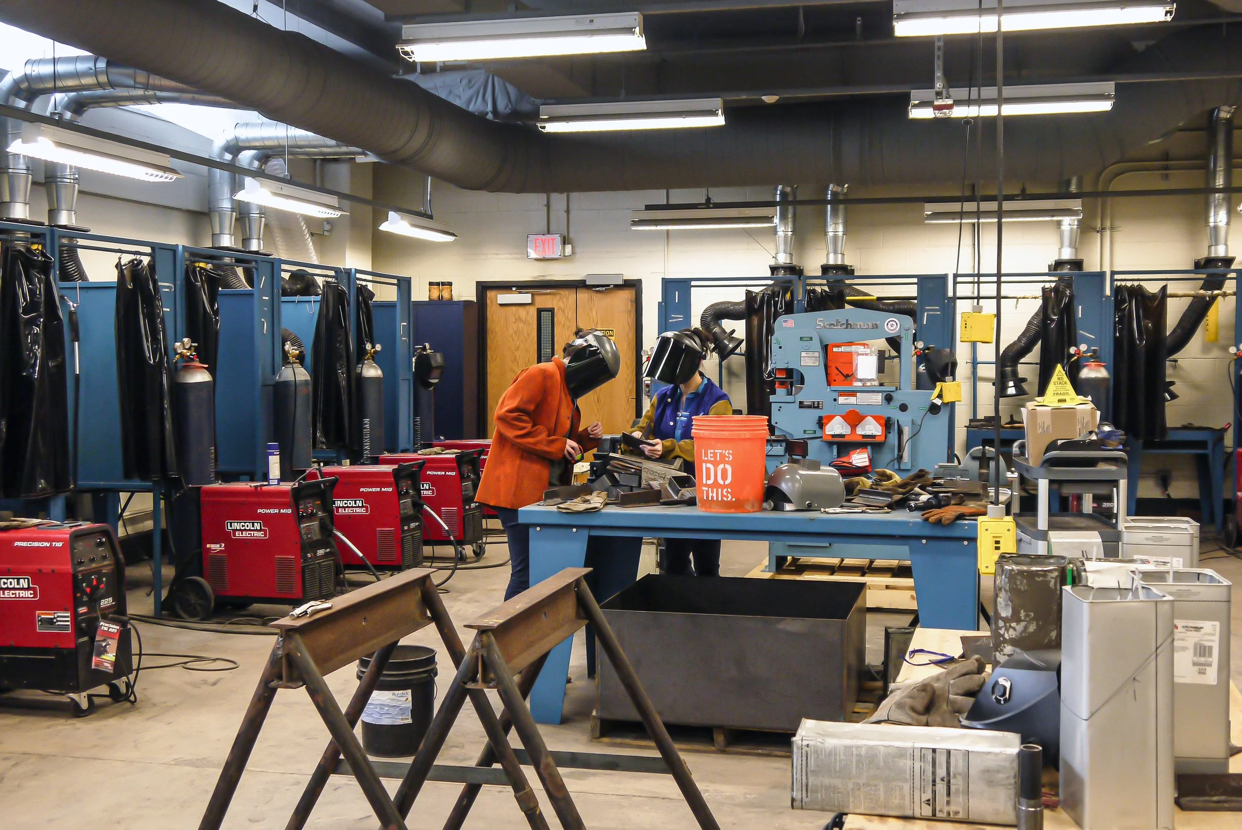 Two workers in a welding shop wearing protective gear, working at a blue workbench with various tools and equipment around them. Welding machines, safety panels, and ventilation ducts are visible in the background.