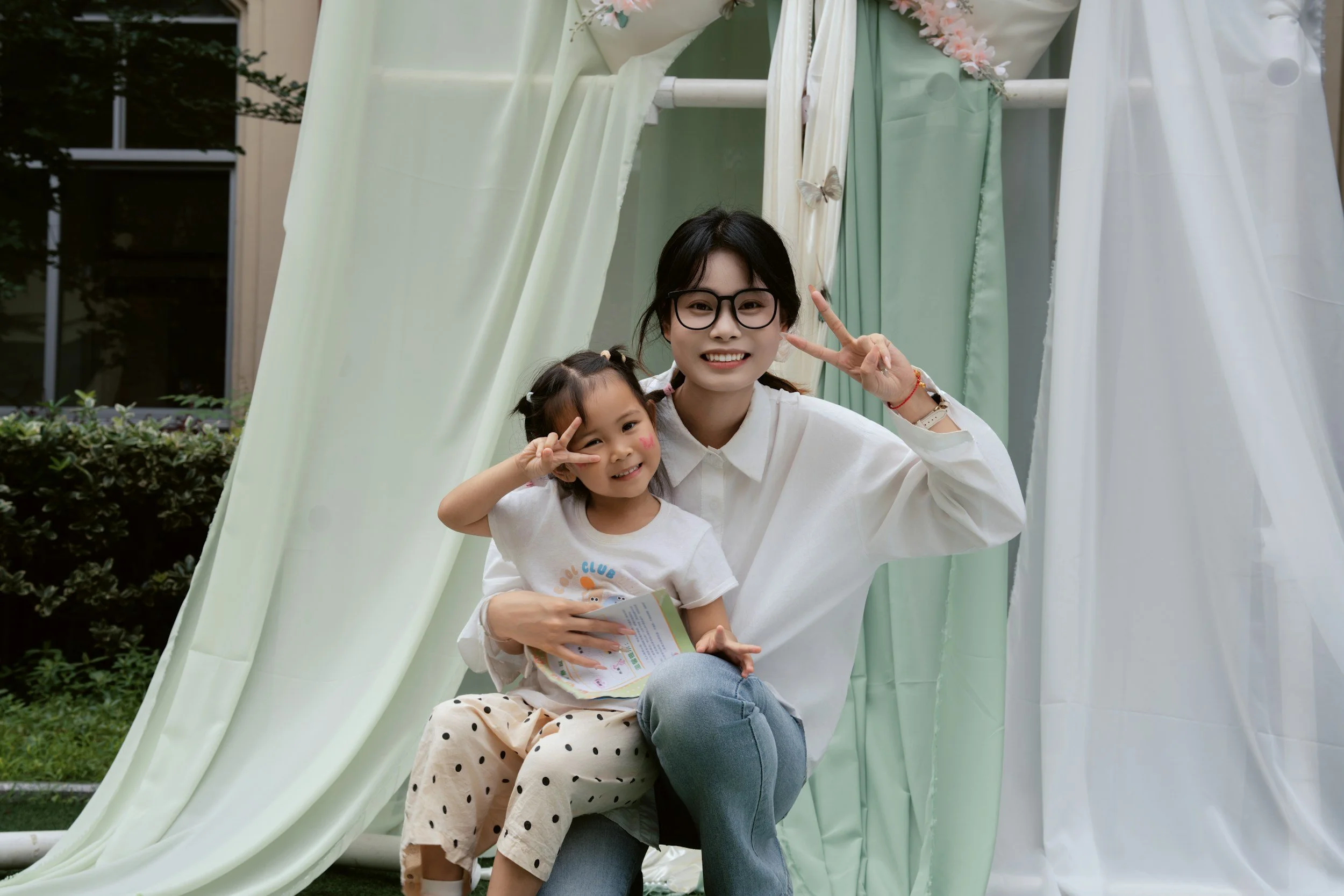 A woman and a young girl sitting inside a fabric fort with light green and white curtains, smiling and making peace signs, outdoors in a garden area.