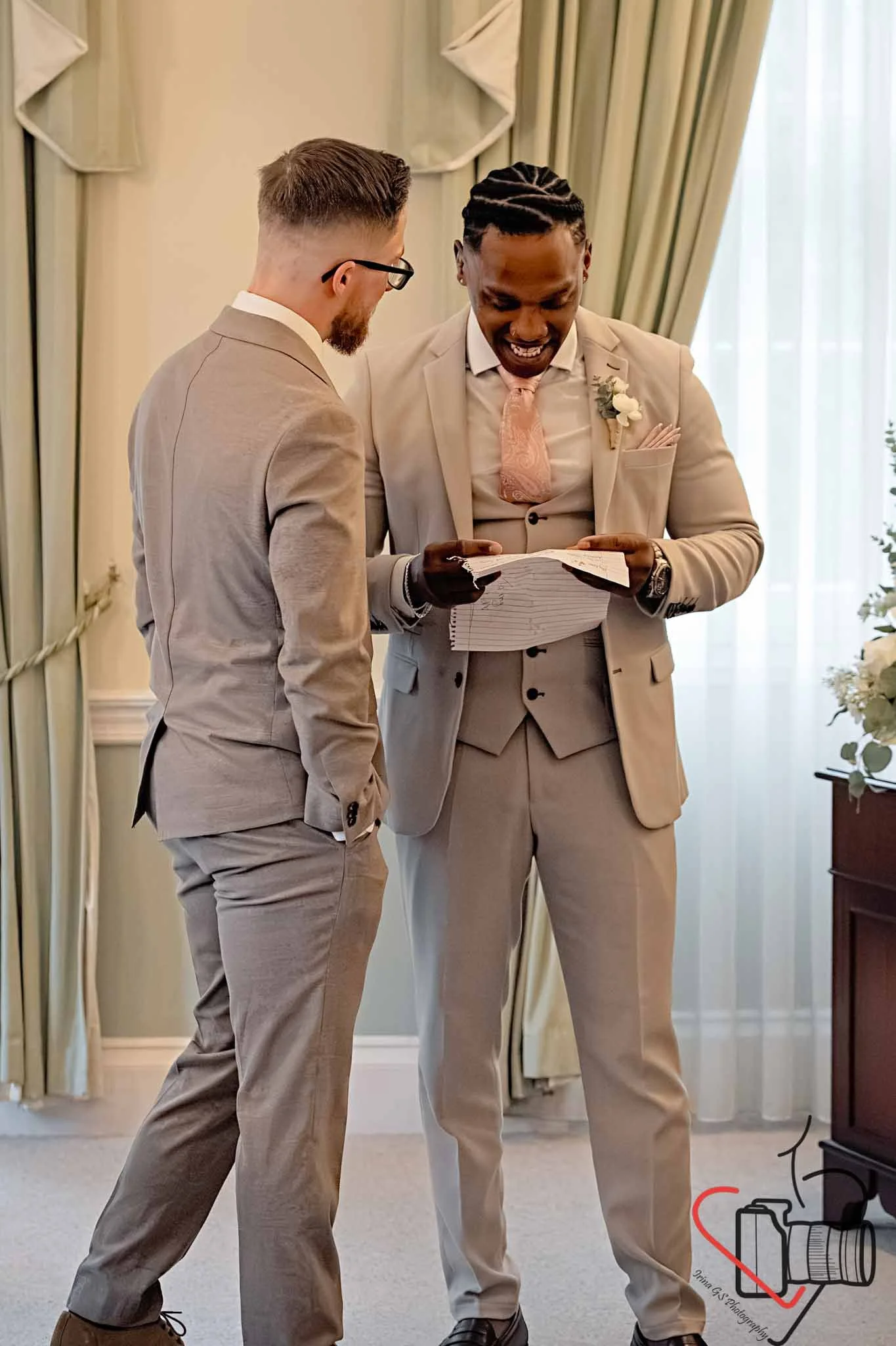 Groom and best men in beige suits are looking at a piece of paper or notes together indoors in a softly lit room with curtains and flowers. Portsmouth Register Office, Milldam House.