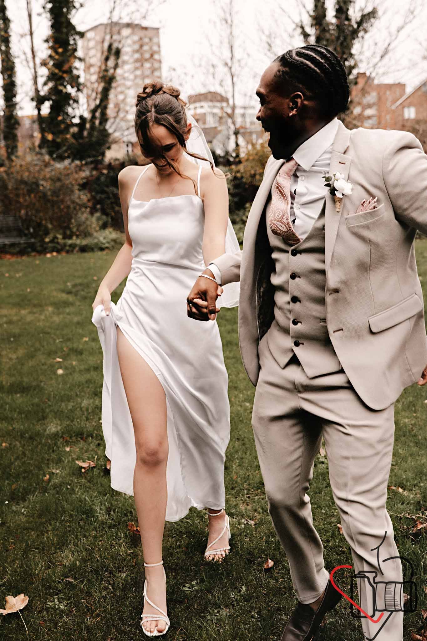 A bride and groom holding hands and smiling outdoors on their wedding day, with trees and buildings in the background. Portsmouth Register Office, Milldam House.