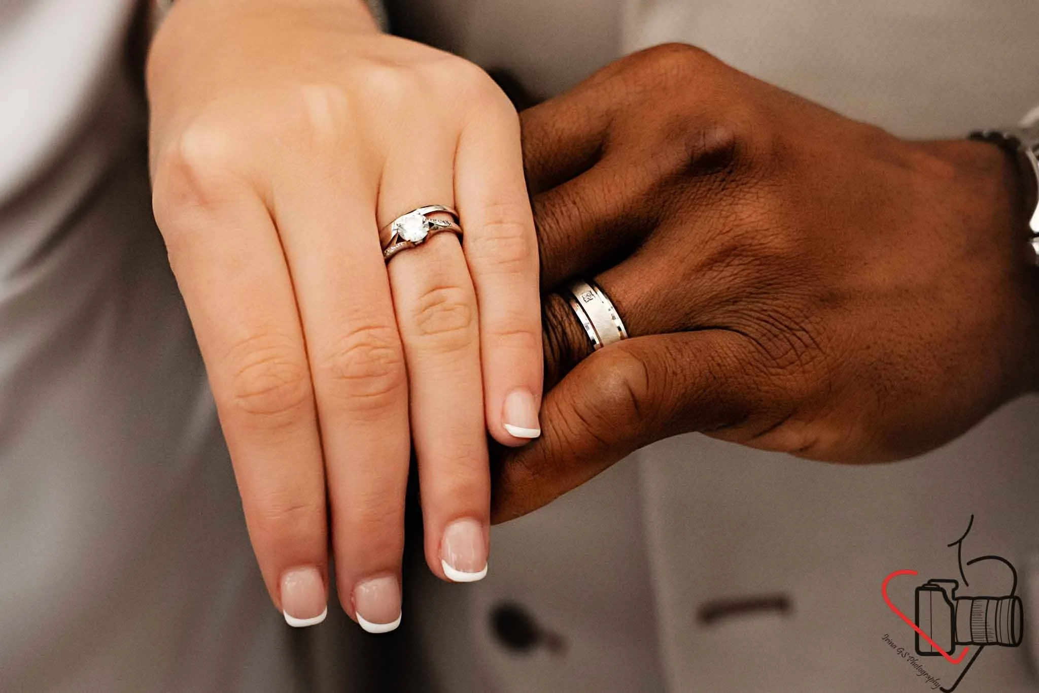 Close-up of a newlywed couple's hands showing wedding rings, with a woman's hand on top of a man's hand. The woman's hand has a diamond engagement ring and a wedding band, and the man's hand has a plain wedding band. The background is blurred.