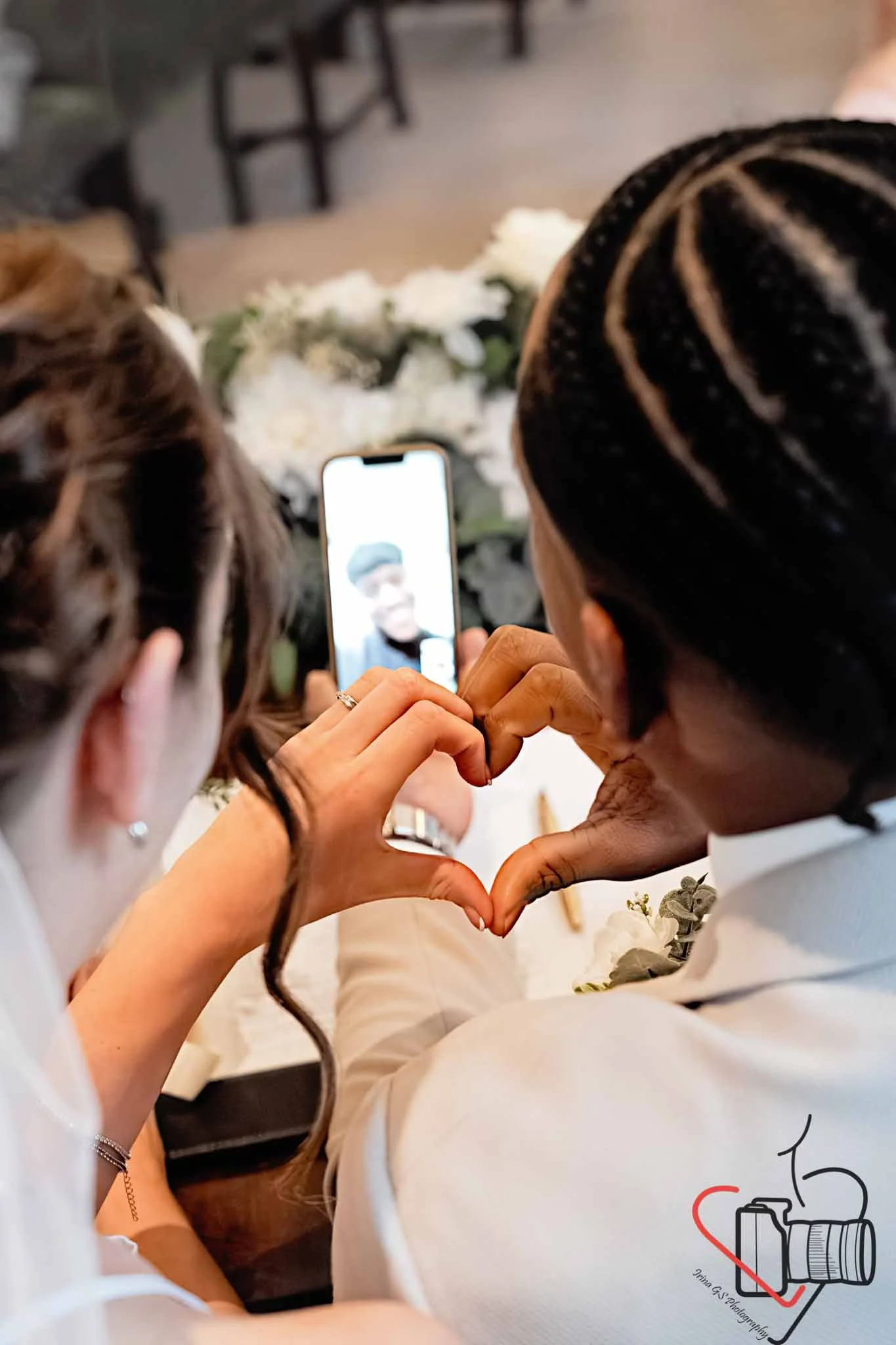 A couple at a wedding sharing a heart shape with their hands while taking a selfie, with a bouquet of white flowers on the table in front of them.