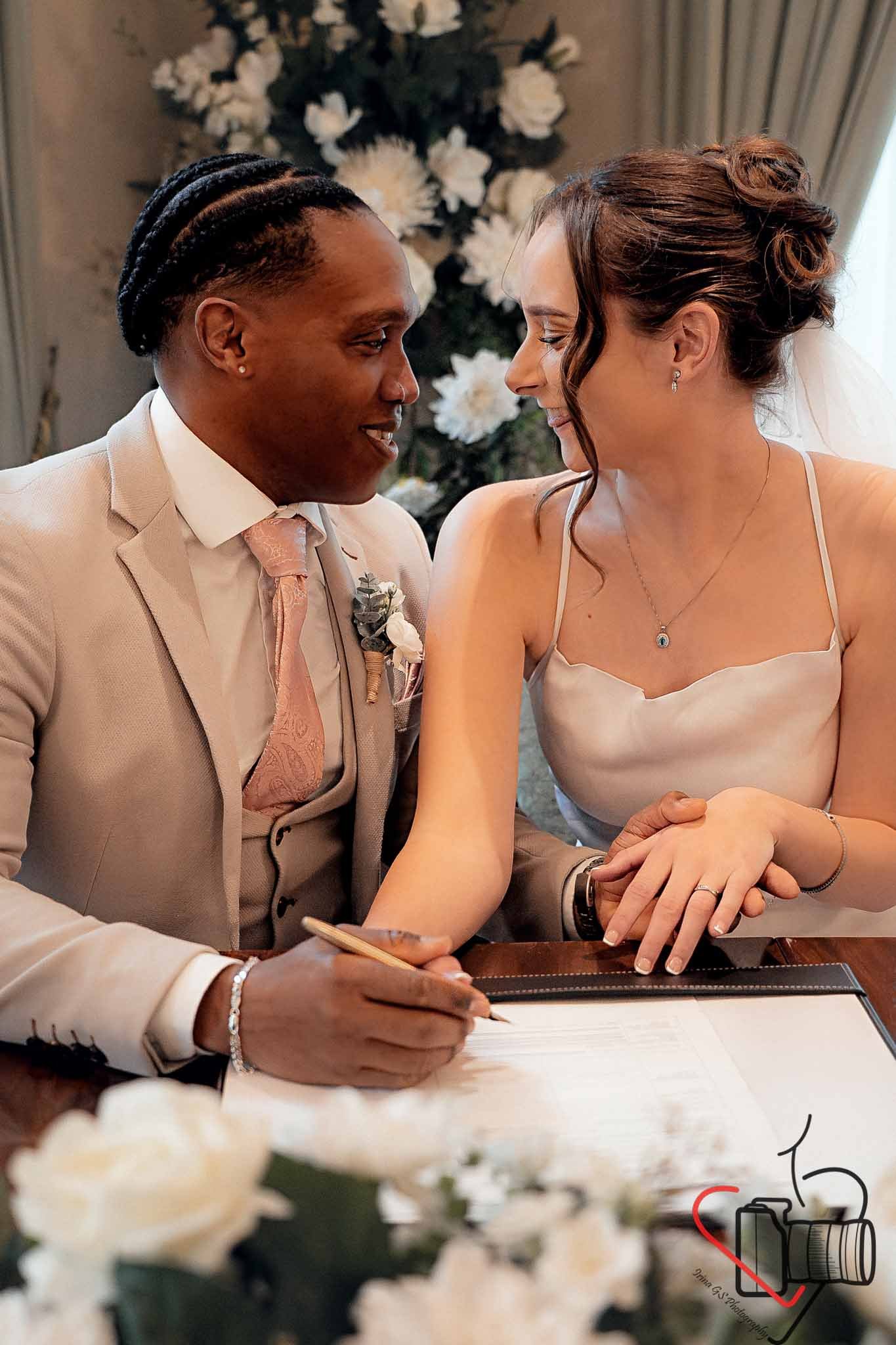A couple gets married, sitting close together, with their foreheads touching. The groom, in a beige suit and tie, is signing a document, while the bride, in a white dress with thin straps and a veil, holds his hand. There are white flowers and a deco