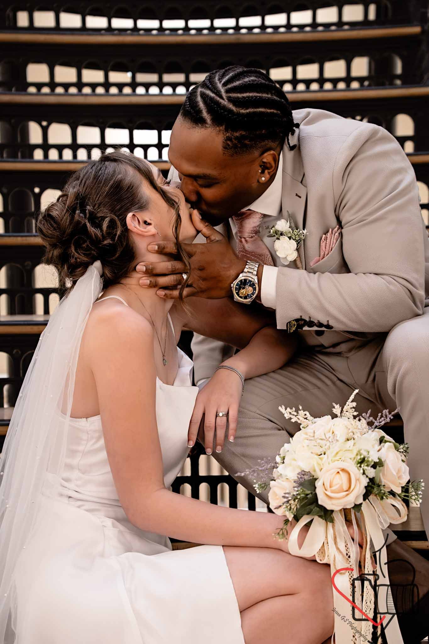 A newlywed couple sharing a kiss during their wedding ceremony, with the bride sitting and holding a bouquet of white and blush roses, while the groom leans in, touching her face. Portsmouth Register Office, Milldam House.
