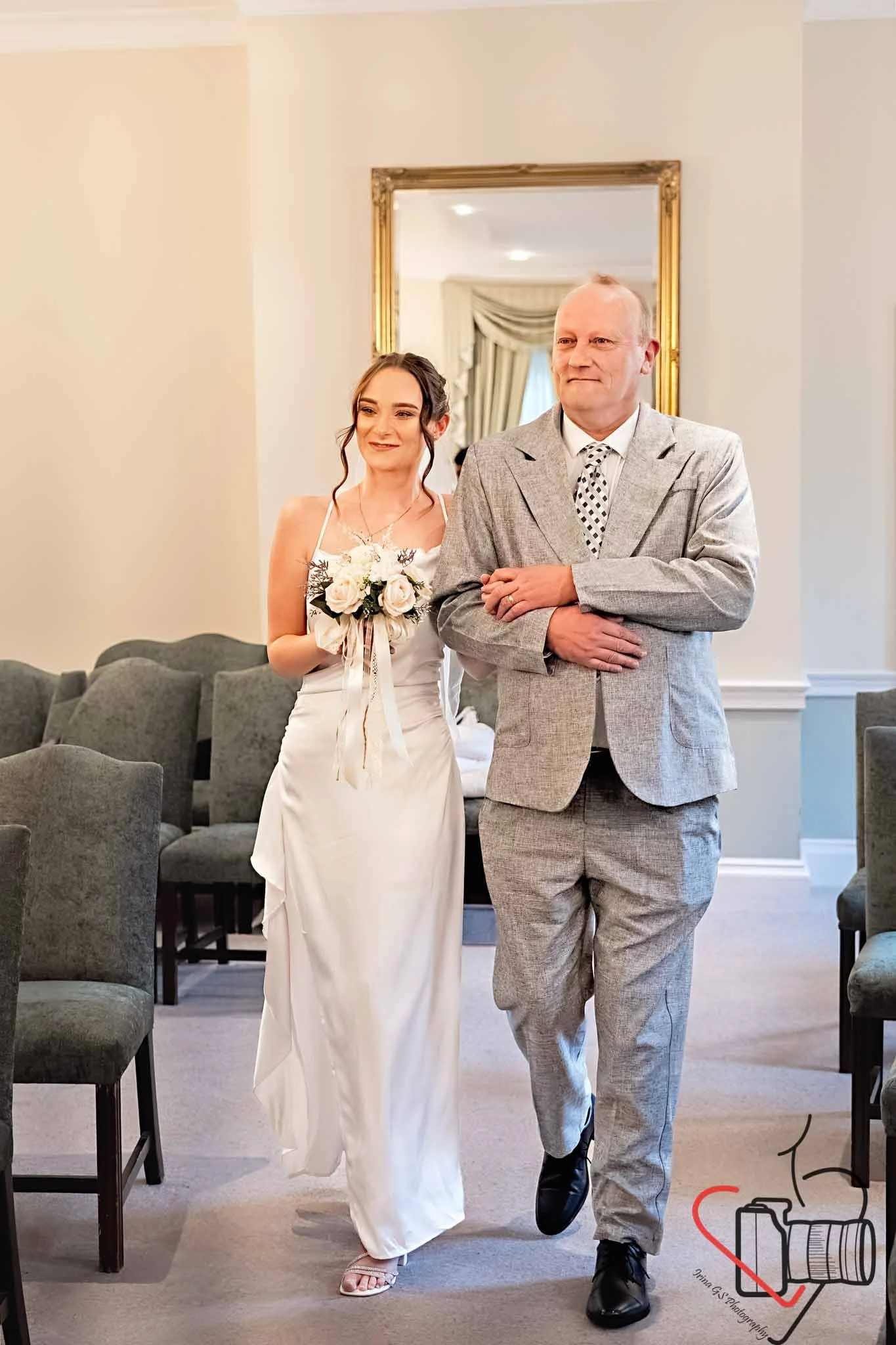 A bride in a white dress holding a bouquet and walking down an aisle, with father in a light gray suit, in a room with beige walls and rows of gray chairs. Portsmouth Register Office, Milldam House.