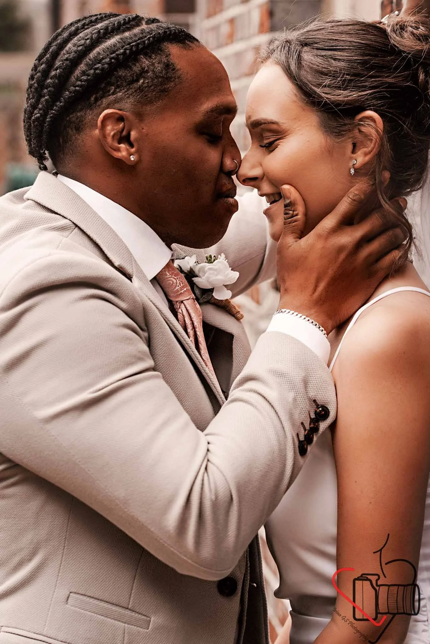 A couple is kissing, with the man gently holding the woman's face. The man is wearing a light-colored suit and the woman is in a white dress. They are close, smiling softly, outdoors near a brick wall. Portsmouth Register Office, Milldam House.