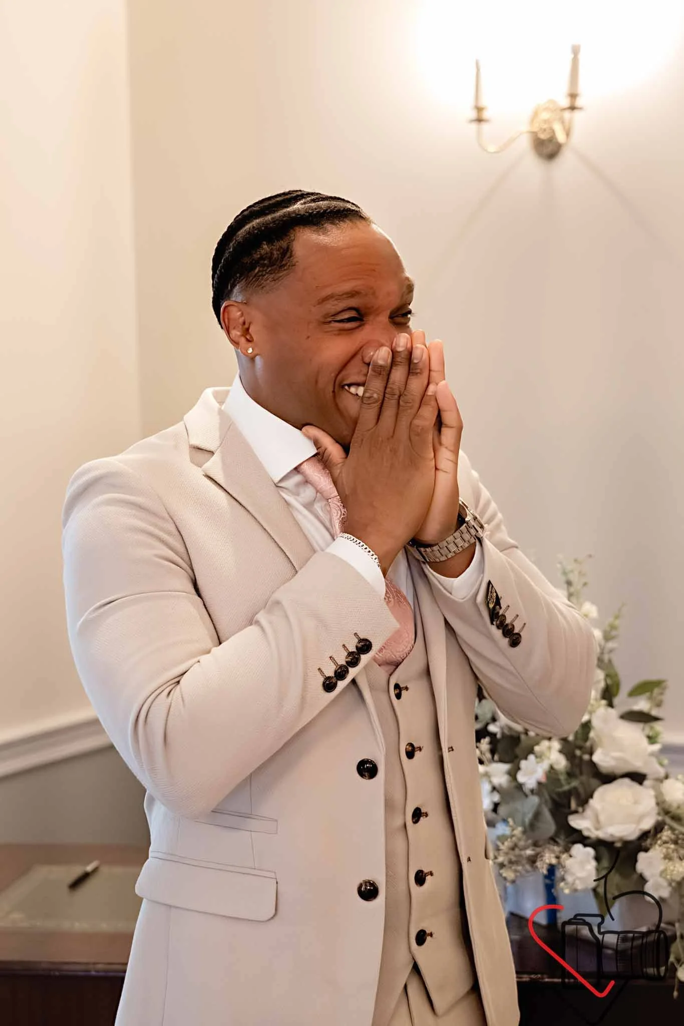 A man in a beige suit covering his face with a smile, standing in front of a white wall with floral decorations. Portsmouth Register Office, Milldam House.
