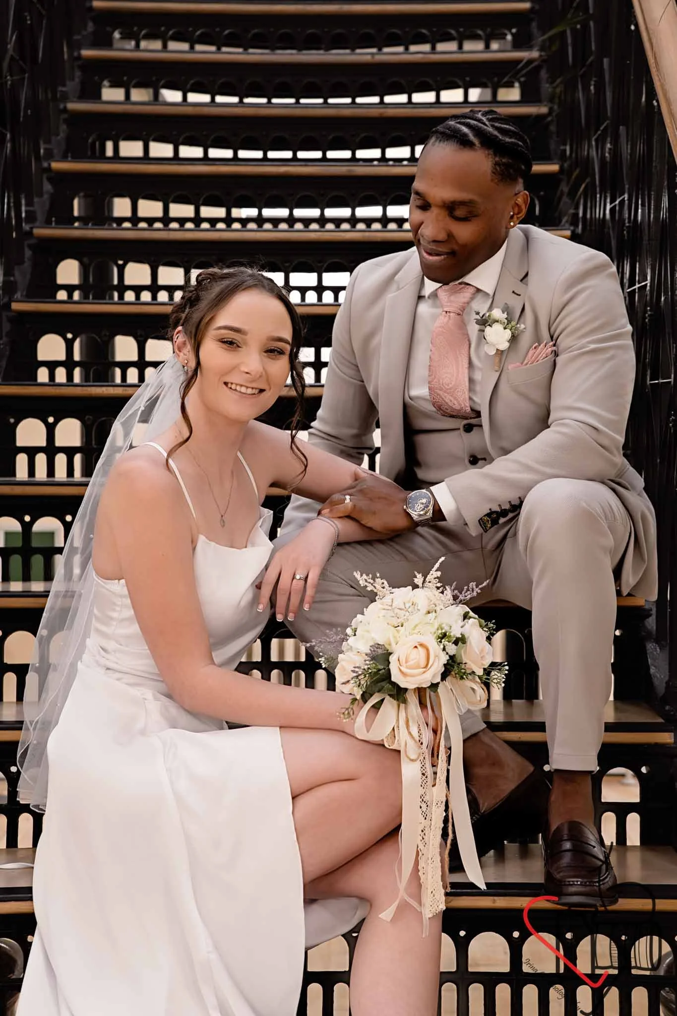 A bride and groom sitting on stairs, with the bride holding a bouquet of flowers, smiling at the camera, and the groom looking at her. Portsmouth Register Office, Milldam House.