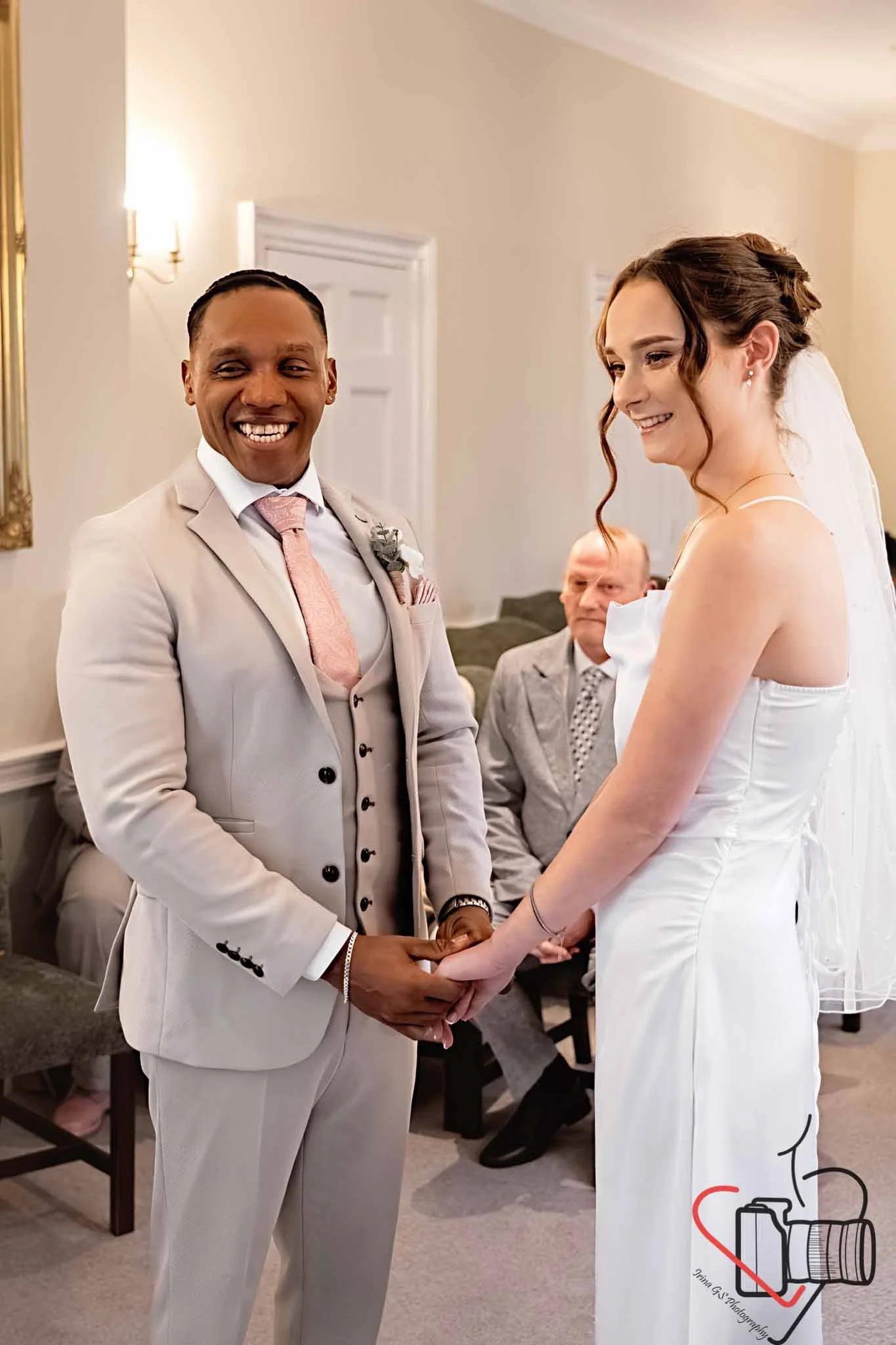 A same-sex couple in wedding attire holding hands during their wedding ceremony, surrounded by seated guests in a decorated room. Portsmouth Register Office, Milldam House.