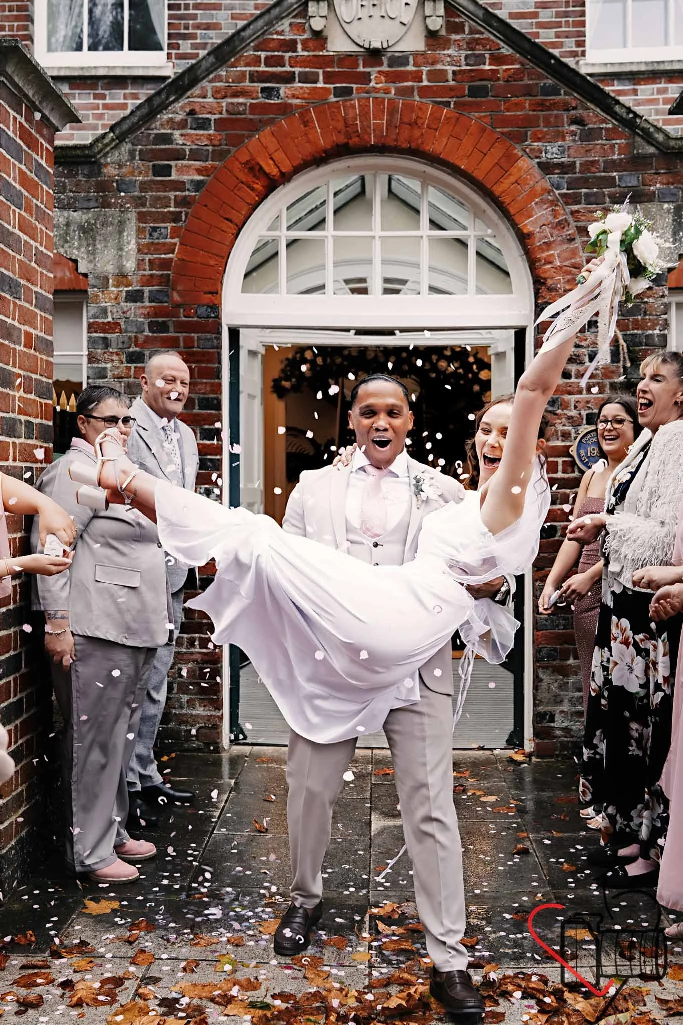 A happy man carrying a woman in a wedding dress, celebrating outside a brick building with guests cheering and confetti falling. Portsmouth Register Office, Milldam House.