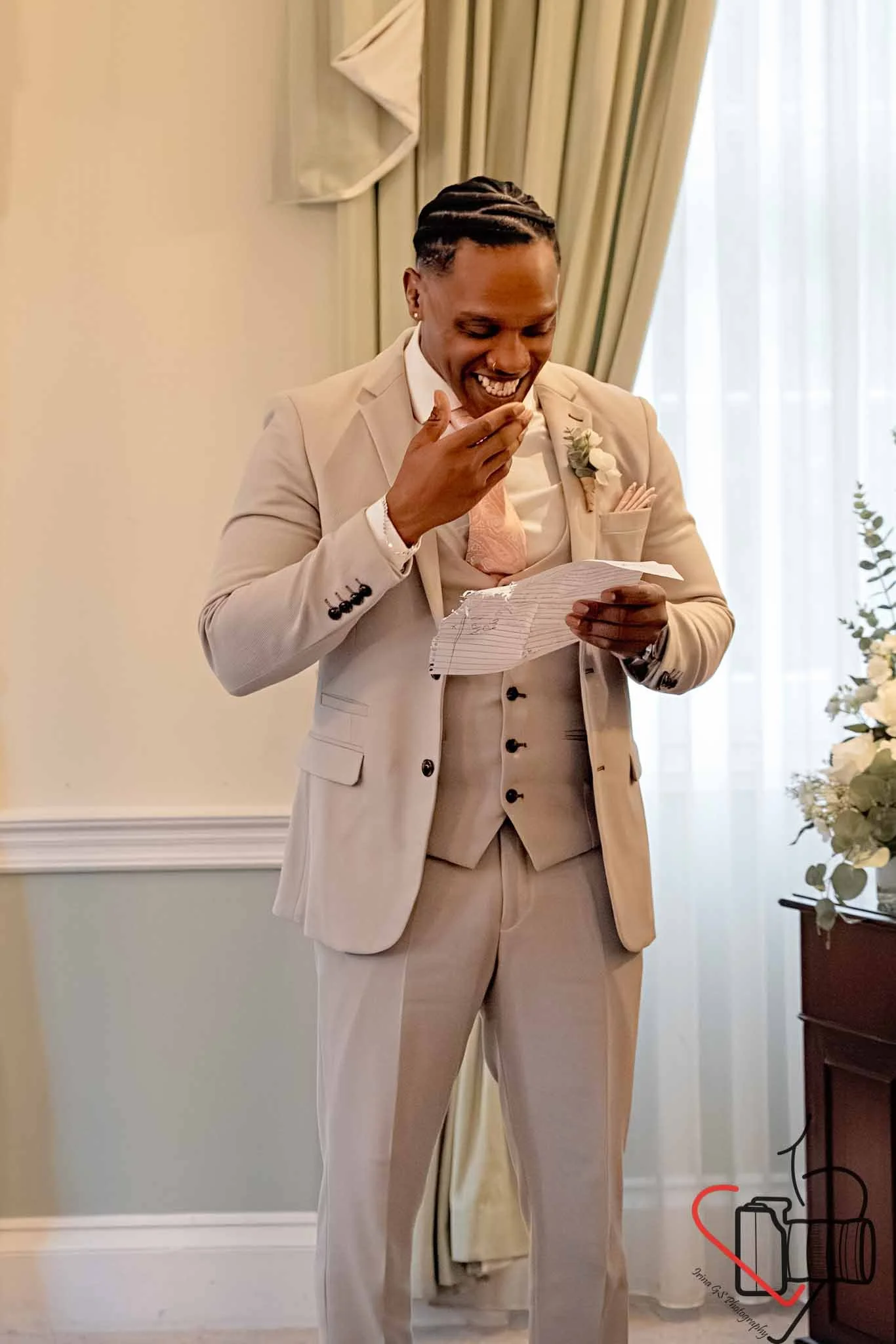 A groom in a beige suit, white shirt, and blush tie, smiling and reading a handwritten note in a well-lit room decorated for a wedding, with flowers and curtains in the background. Portsmouth Register Office, Milldam House.