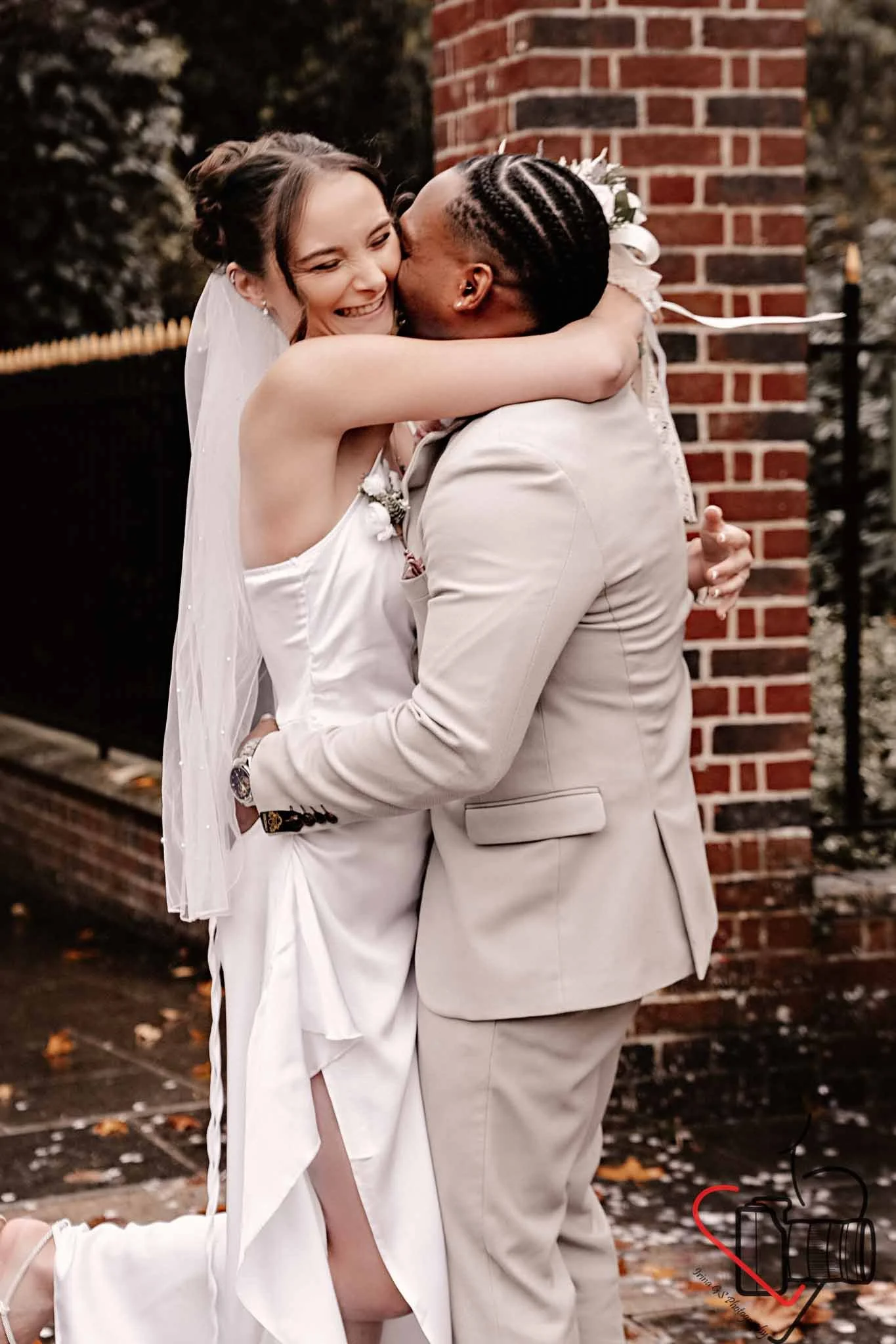 A bride and groom sharing a hug outside near a brick wall, dressed in wedding attire. The bride is smiling with her arms around the groom's neck, and the groom is holding the bride up while kissing her on the cheek. Portsmouth Register Office