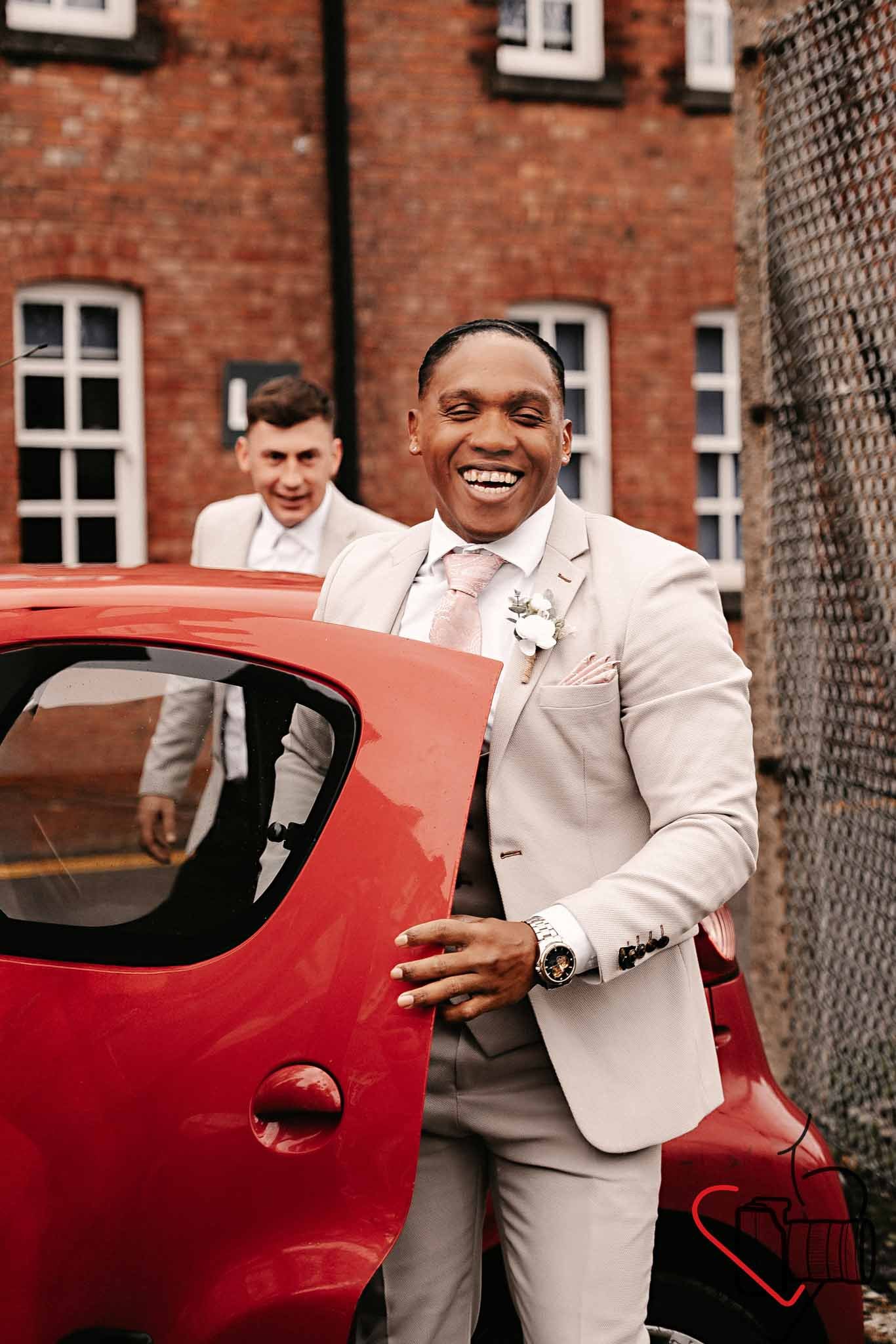 Two men in light-colored suits, one in the foreground and one in the background, are smiling and standing next to a red car outdoors with brick buildings in the background. Portsmouth Register Office, Milldam House.