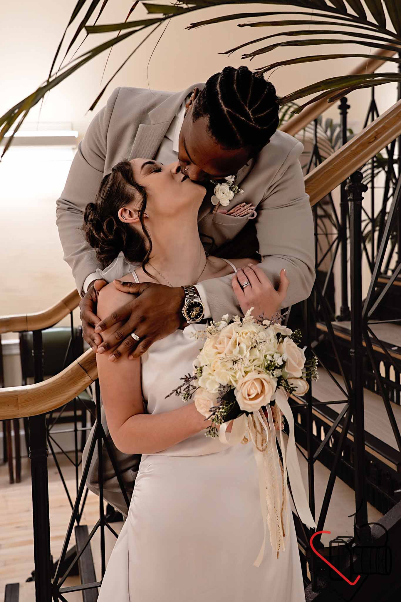 A newlywed couple sharing a romantic kiss on a staircase, with the bride holding a bouquet of white and peach roses. Portsmouth Register Office, Milldam House.