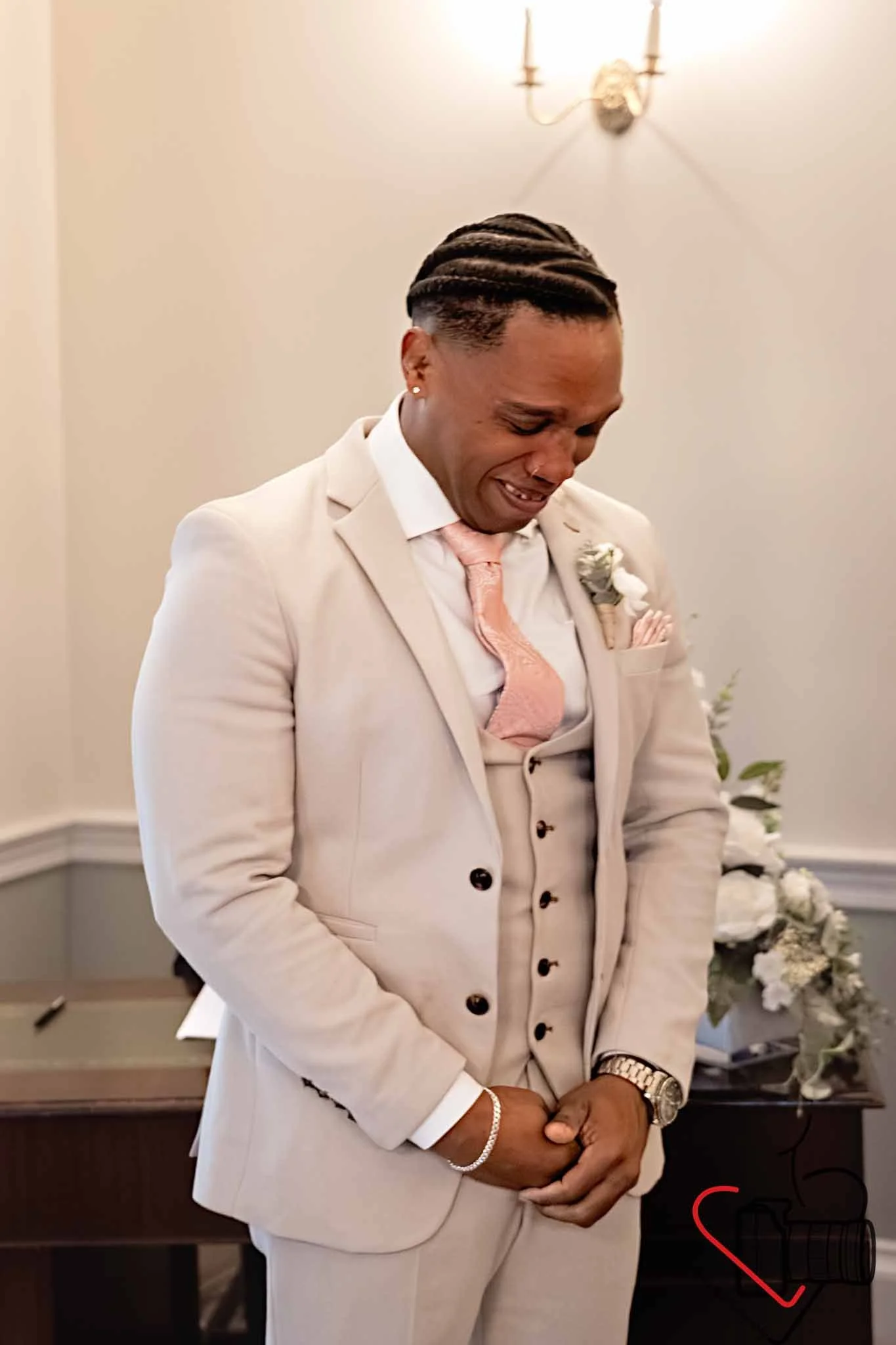 A man dressed in a beige suit with a pink tie and boutonniere is smiling and standing with his eyes closed in a room with a wall-mounted light fixture and a table with flowers in the background. Portsmouth Register Office, Milldam House.