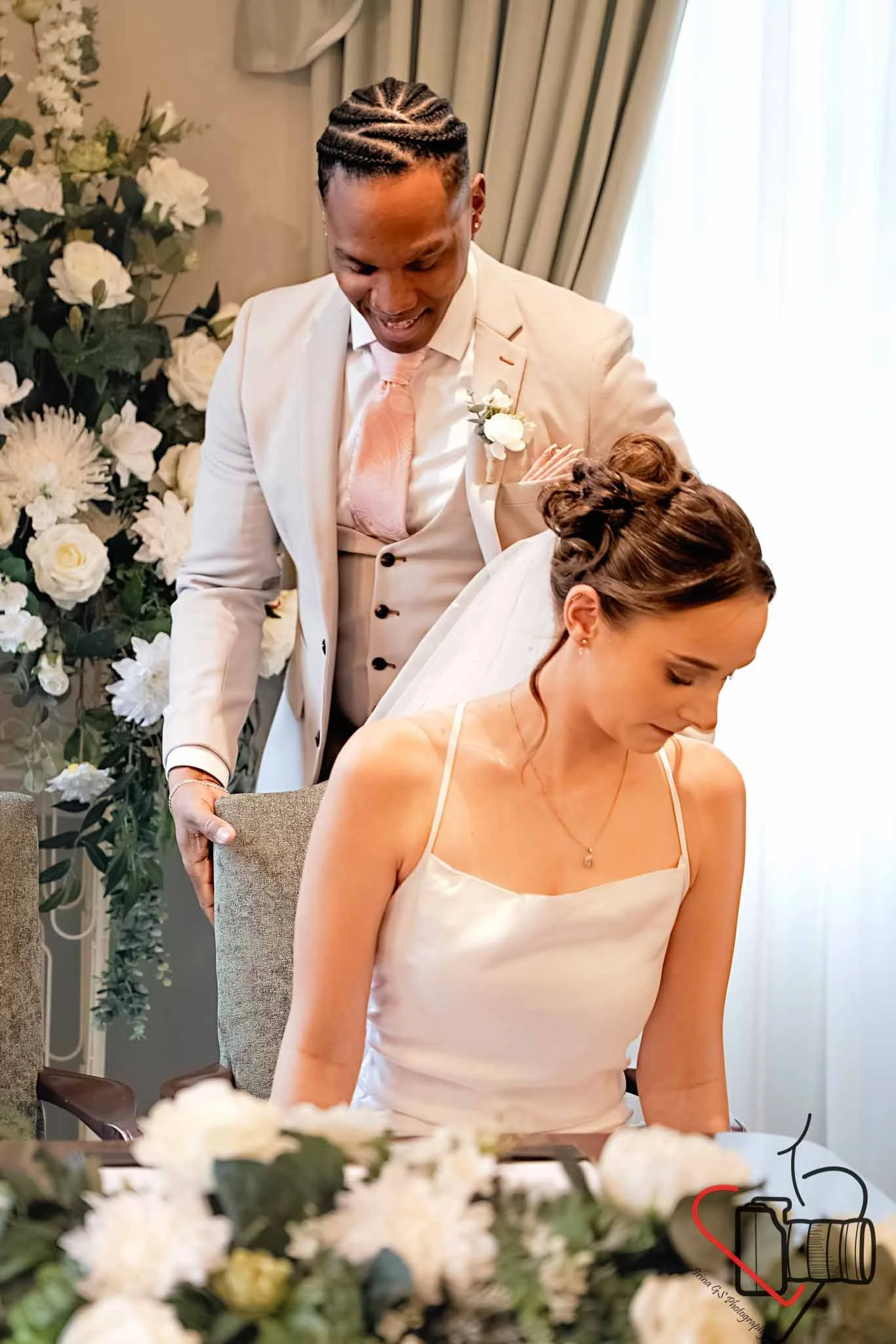 A groom in a beige suit with a pink tie and a white boutonniere stands behind a seated bride, who is in a spaghetti strap white dress with her hair in an updo, during a wedding ceremony in a decorated room with white flowers and curtains. 