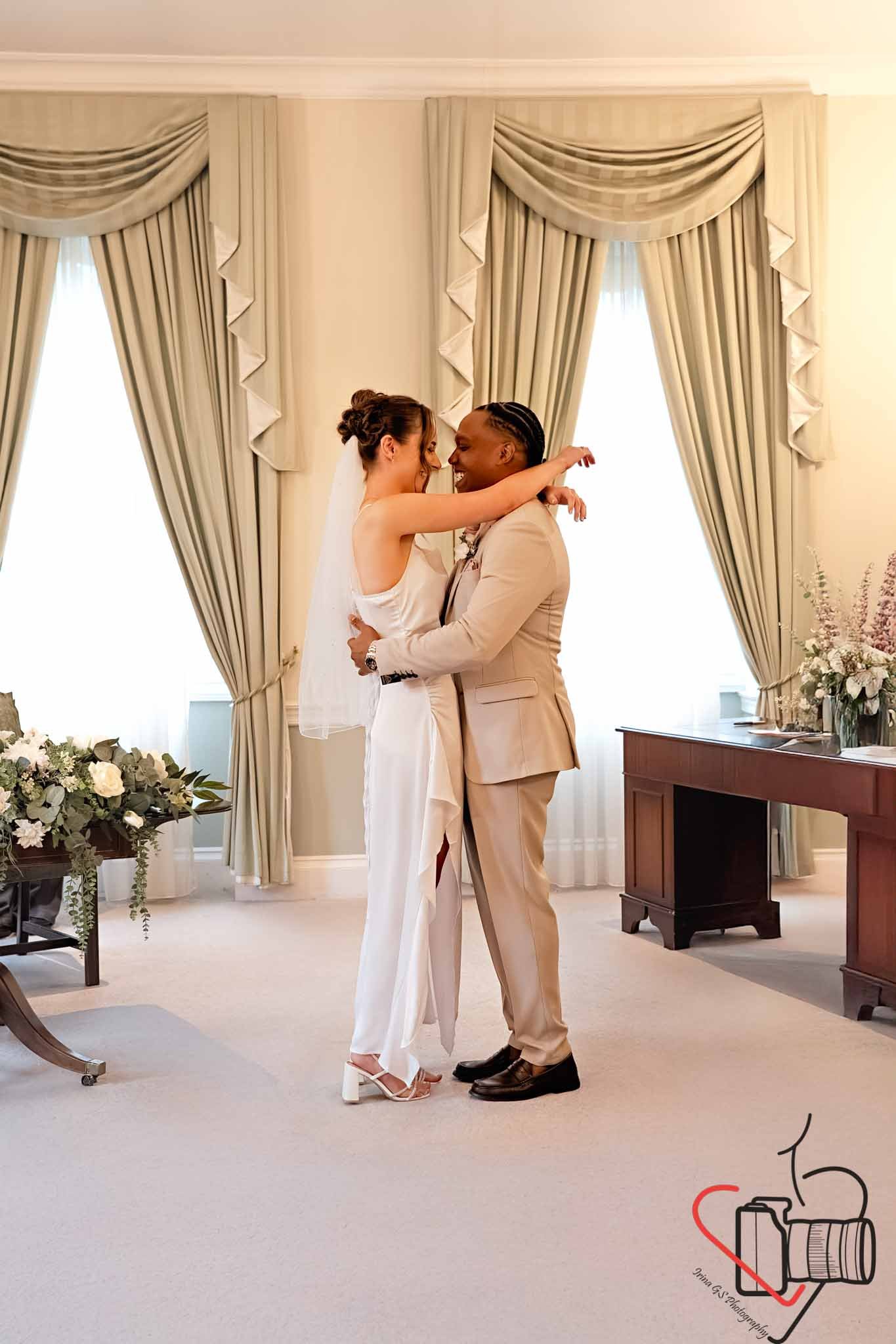 A couple in wedding attire embracing and smiling in a room with cream-colored walls, large windows with draped curtains, and floral arrangements. Portsmouth Register Office, Milldam House.