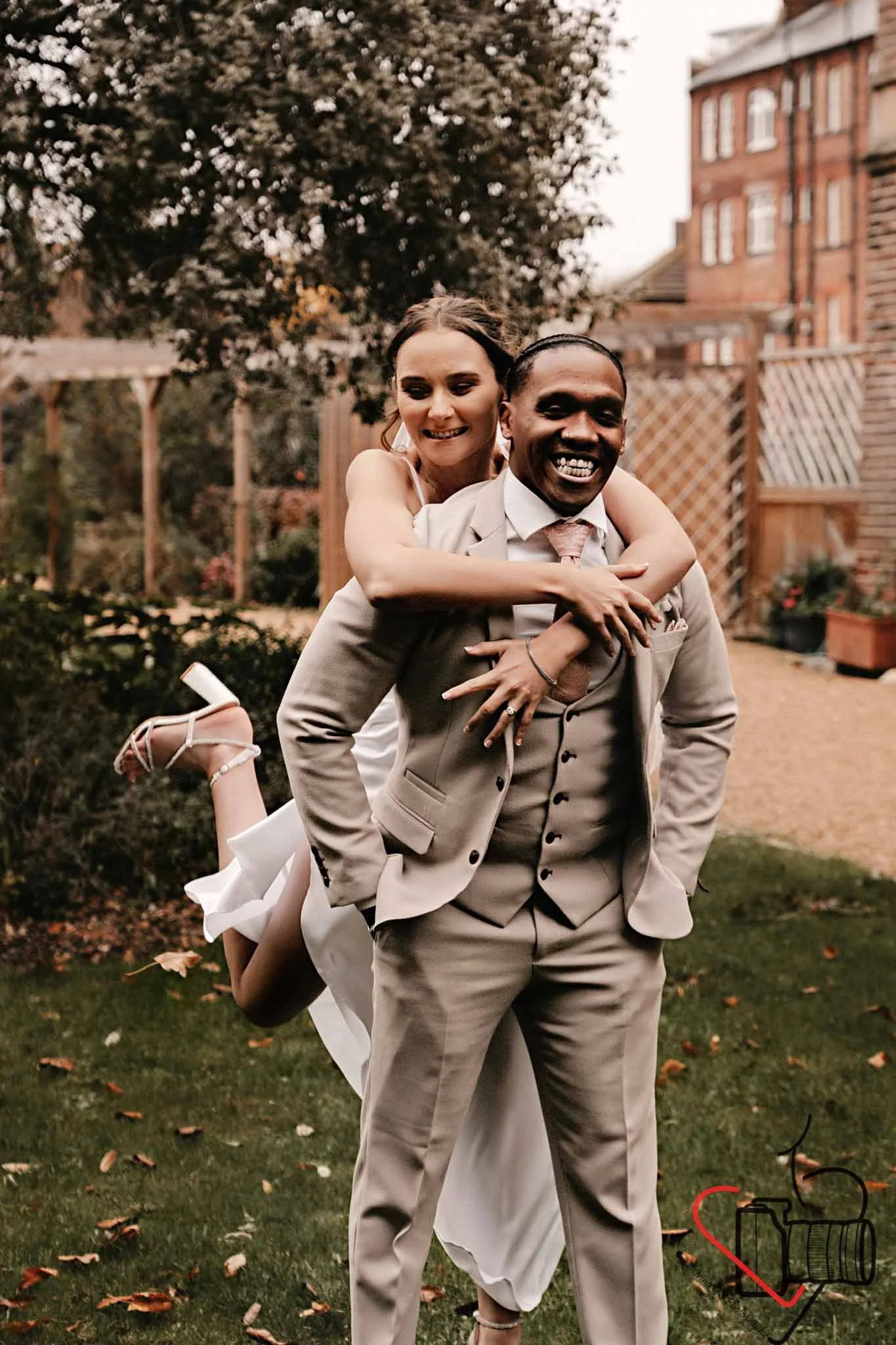A smiling bride piggyback riding on a groom's shoulders outdoors, with trees and a red brick building in the background. Portsmouth Register Office, Milldam House.