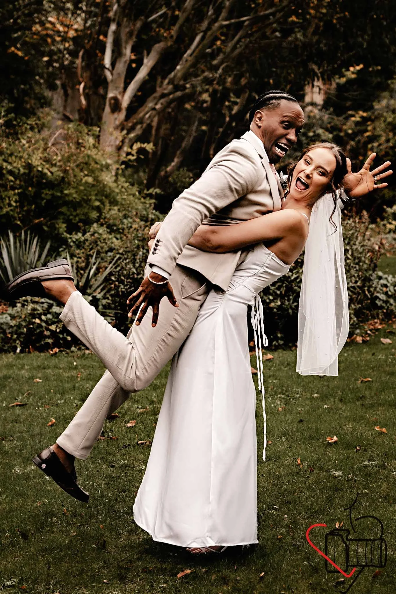 A joyful couple in wedding attire playfully lift each other outdoors on a grassy area with trees and bushes in the background, smiling and laughing. Portsmouth Register Office, Milldam House.