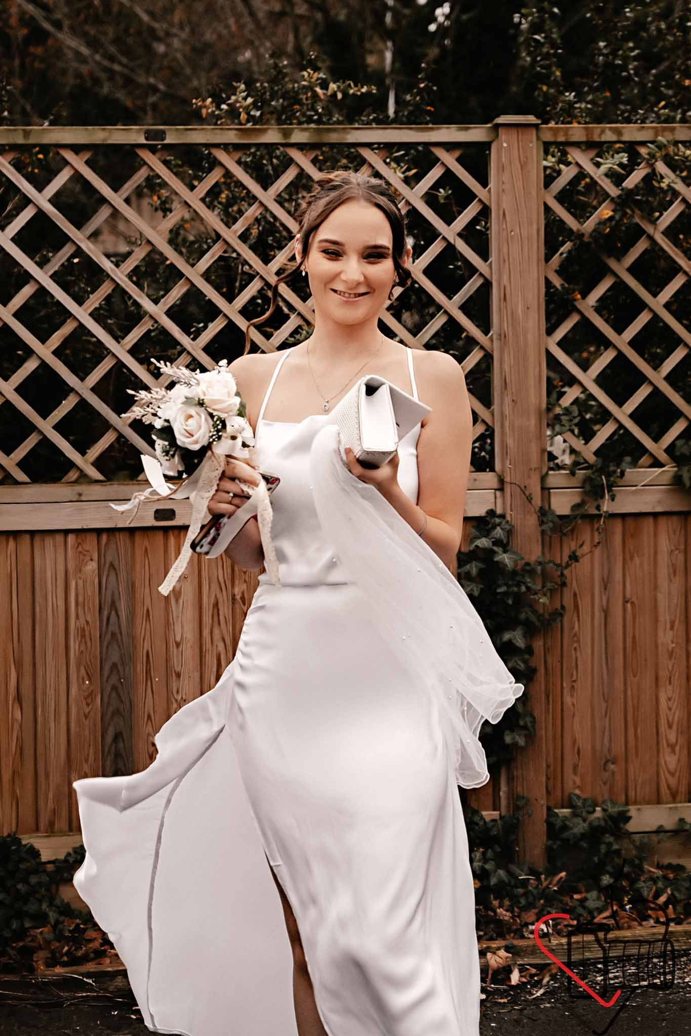 A woman in a white wedding dress holding a bouquet of flowers and a small purse, smiling, standing outdoors in front of a wooden fence.