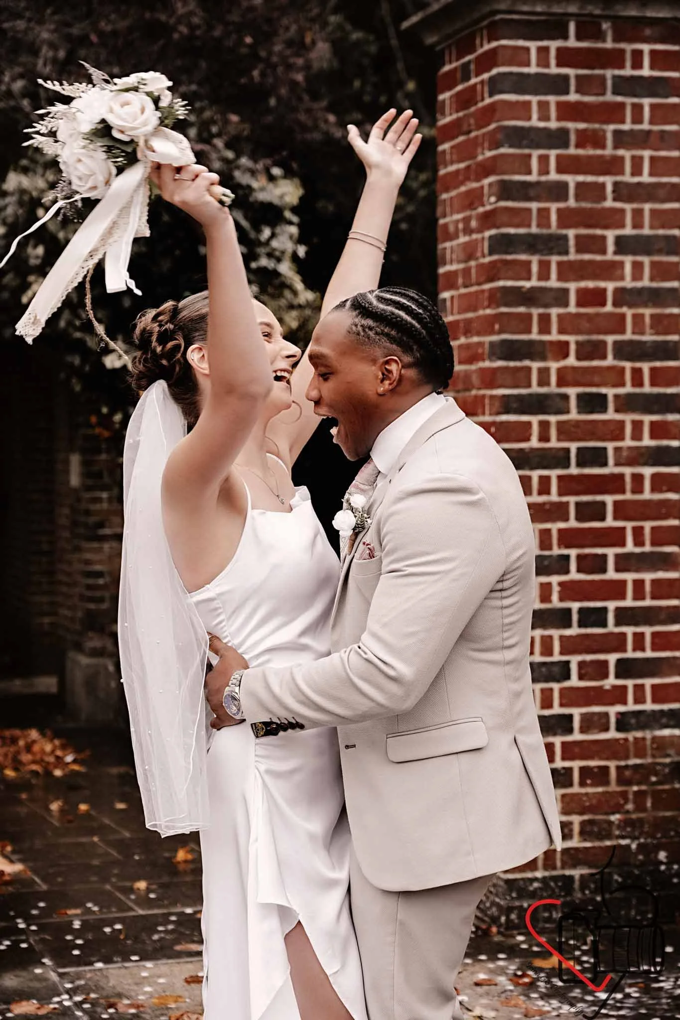A bride and groom are celebrating their wedding outdoors near a brick wall. The bride, in a white wedding dress and veil, is holding a bouquet above her head and laughing. The groom, in a light-colored suit, is smiling and holding the bride.