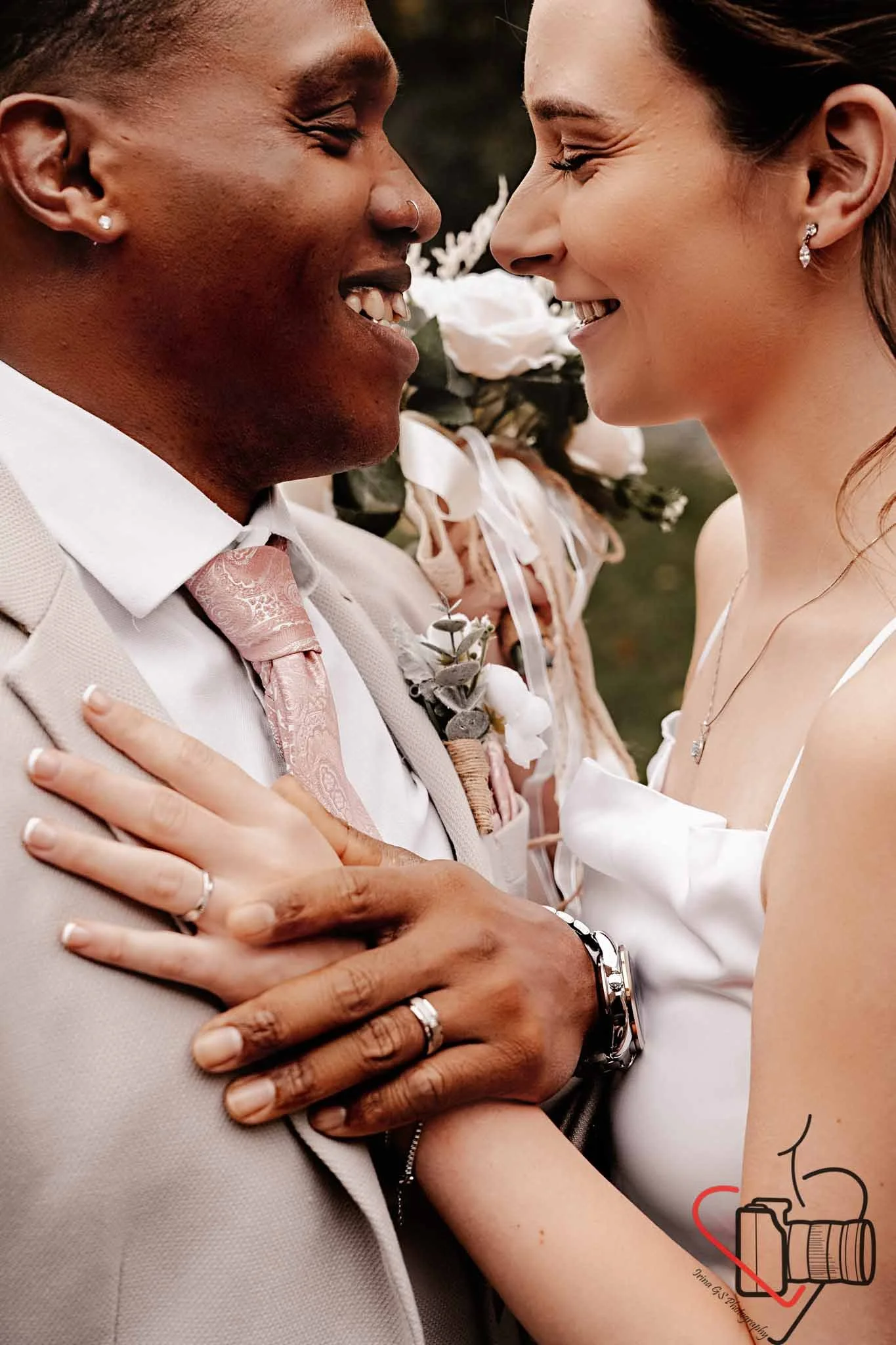 Close-up of a smiling couple at their wedding, gazing into each other's eyes, with the bride holding the groom's hands, and flowers in the background.
