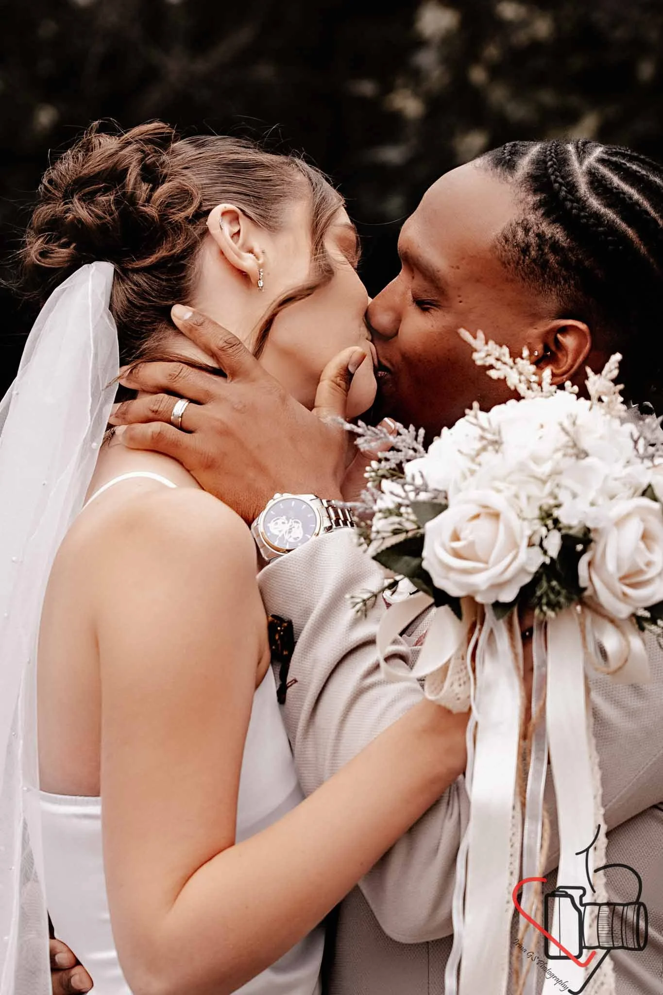 A newly married couple sharing a passionate kiss, with the bride holding a bouquet of white flowers and wearing a white wedding dress and veil, and the groom in a beige suit with a smartwatch on his wrist.