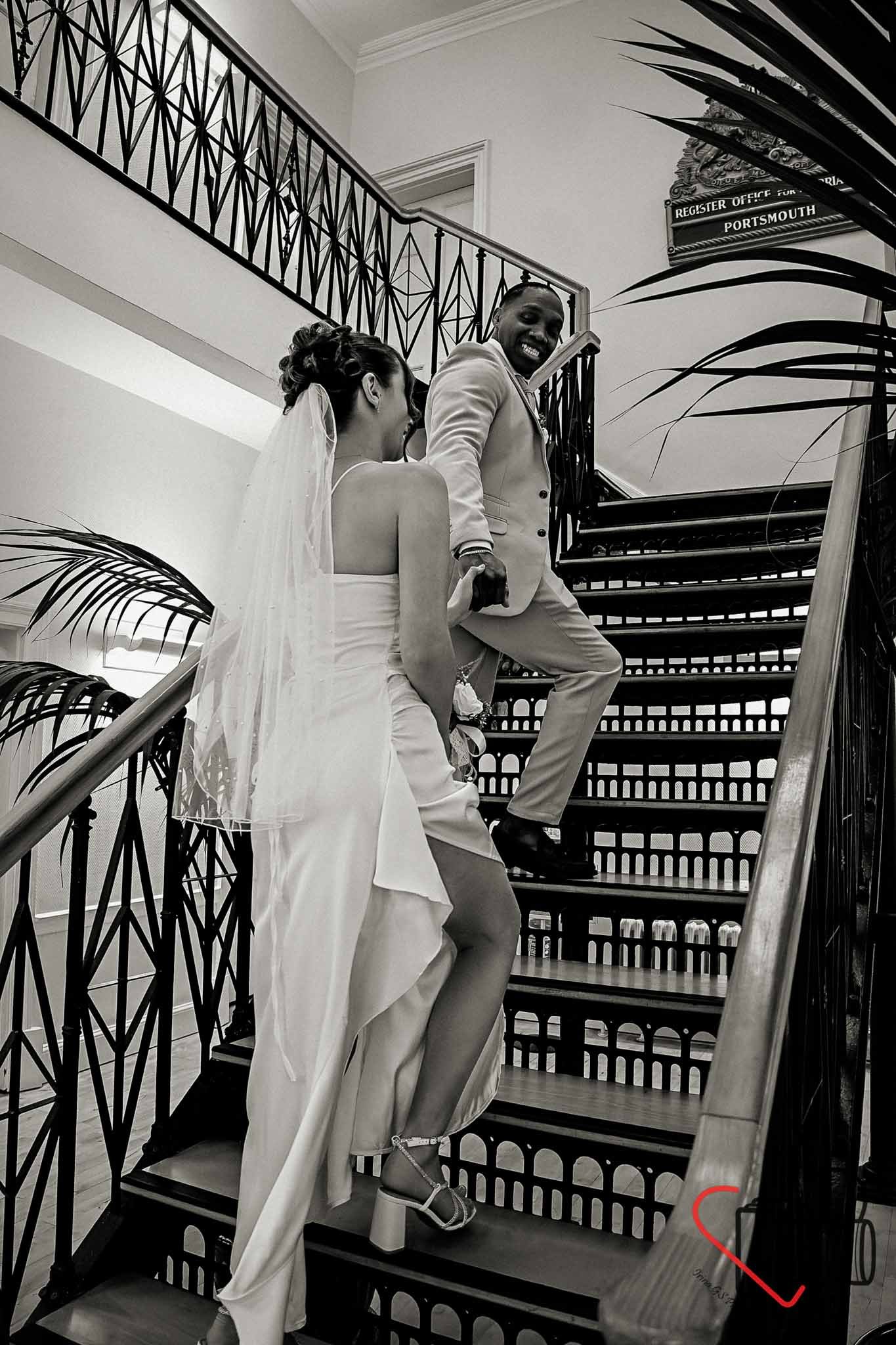 A bride and groom in wedding attire on a staircase, smiling and looking at each other, with the groom standing on the stairs and the bride sitting on the steps. Portsmouth Register Office, Milldam House.