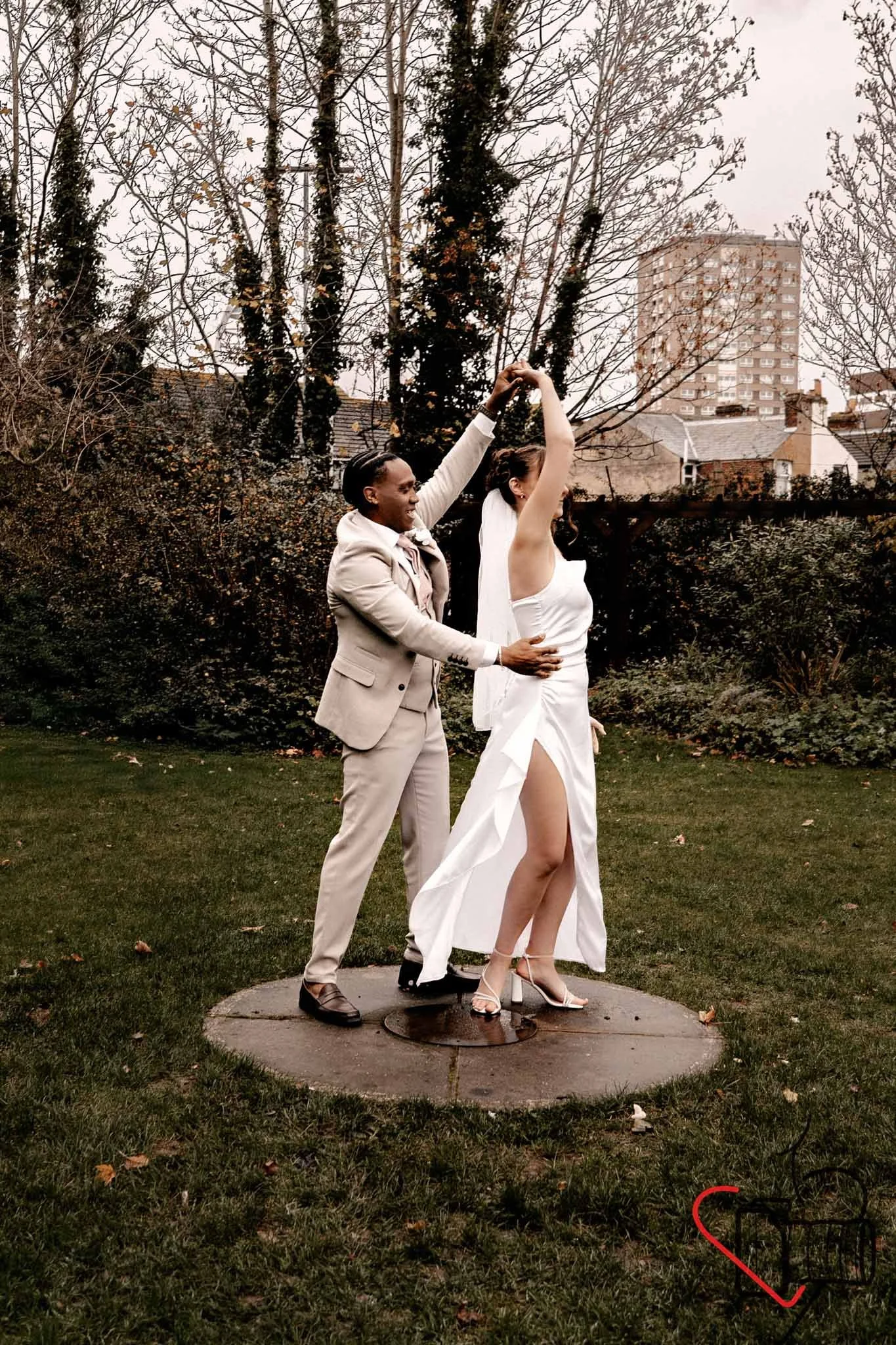 A couple is dancing outside in a park, with tall trees and a building in the background. The woman is wearing a white dress, and the man is in a beige suit. They are holding hands, mid-dance, on a circular concrete platform.Portsmouth Register Office