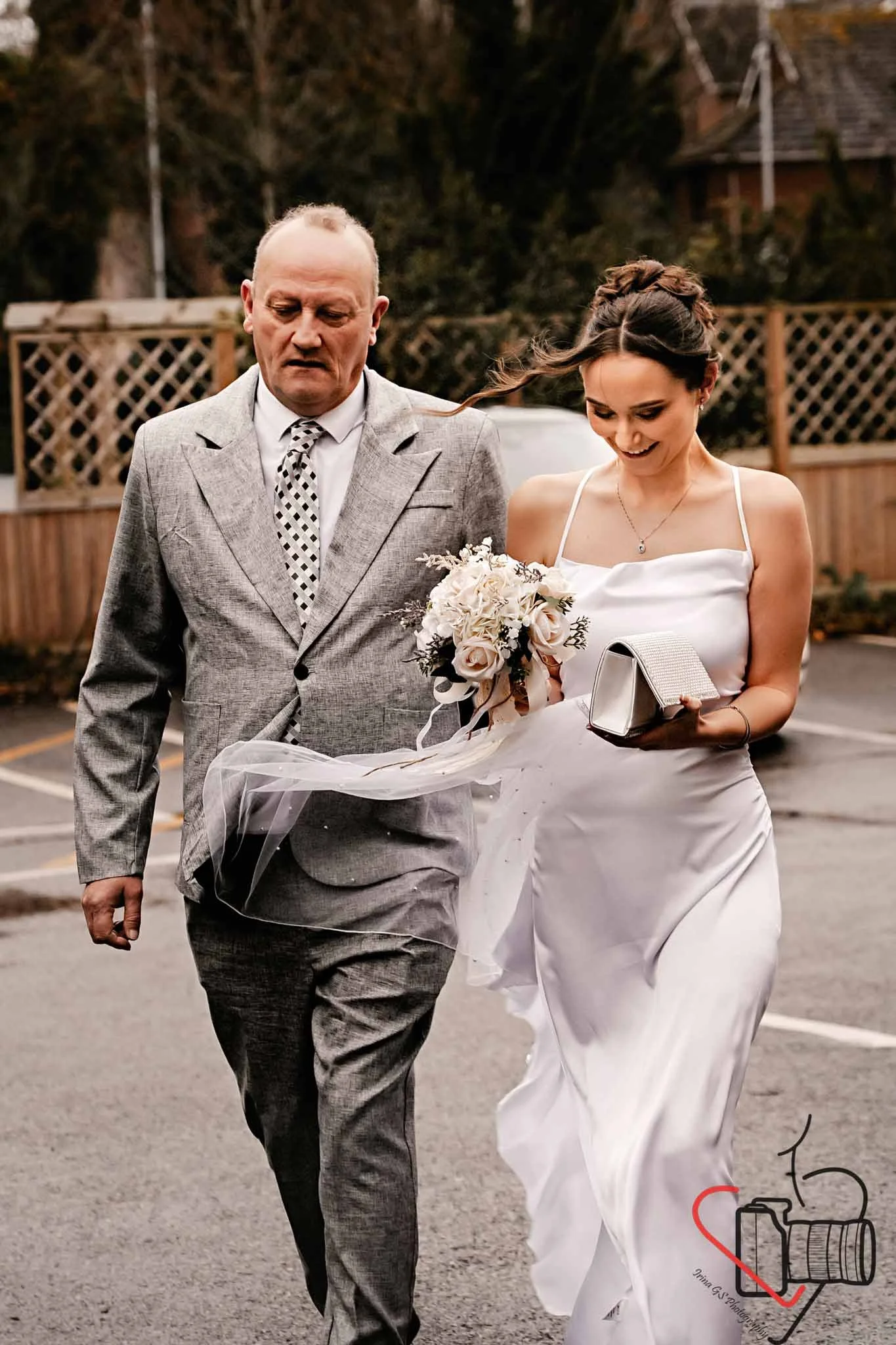 A bride and an older man walking together outdoors on a gray paved surface, with the bride holding a bouquet and an elegant clutch, and the father wearing a gray suit with a patterned tie. A wooden fence and trees are visible in the background.