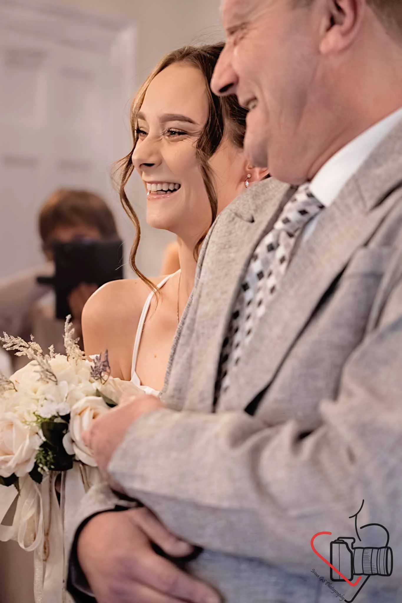 A woman and man smiling during a wedding ceremony. The woman is holding a bouquet of flowers and the man has his arm around her. The background shows other people attending the event.