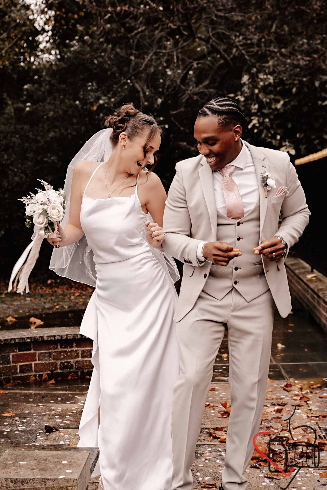 A bride and groom are celebrating outdoors, smiling and holding hands, with the bride wearing a white wedding dress and holding a bouquet, and the groom in a light-colored suit with a pink tie. Portsmouth Register Office, Milldam House.