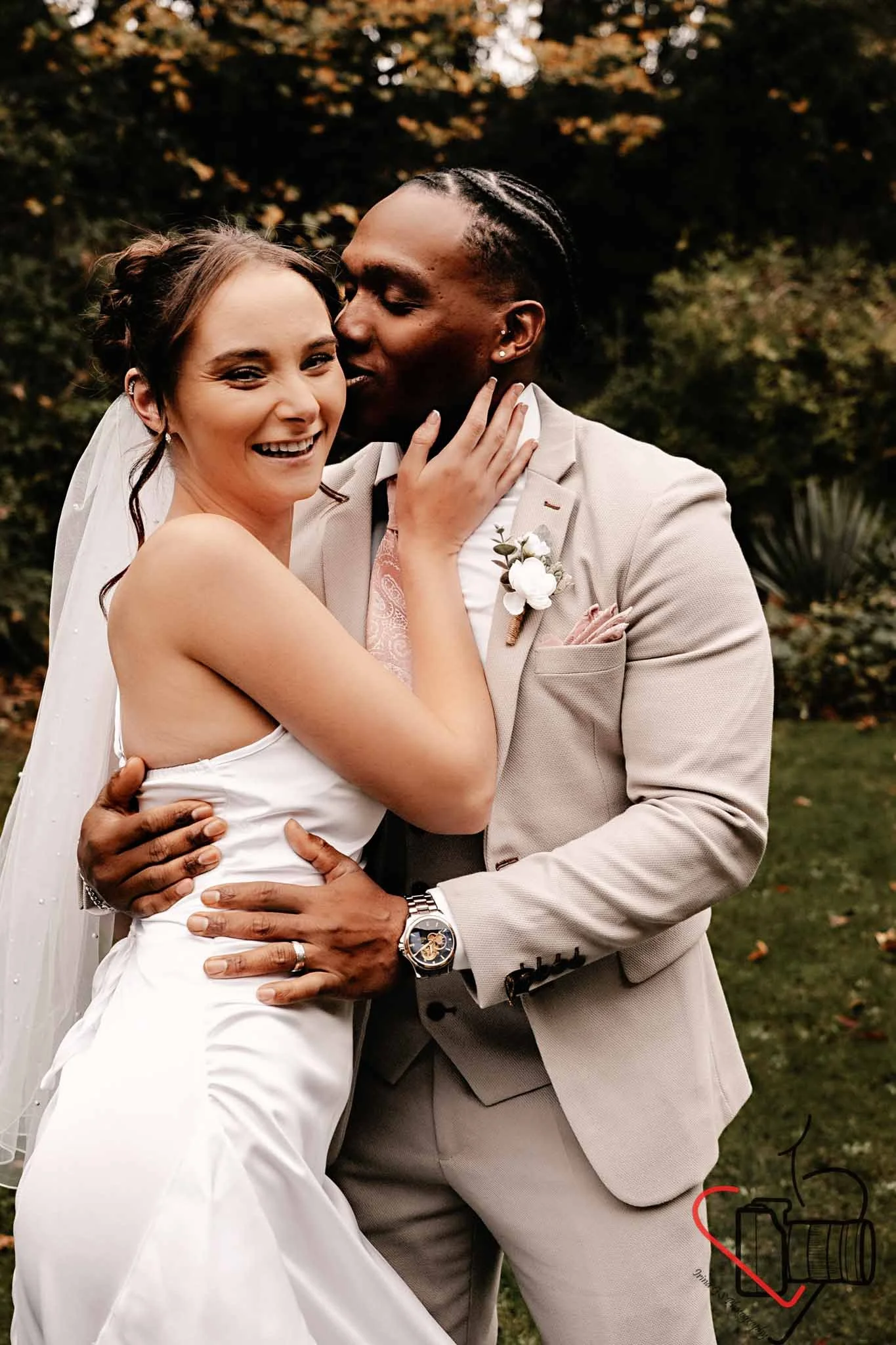 A newlywed couple sharing a loving embrace outdoors, with the groom kissing the bride's cheek. The bride is smiling and wearing a white wedding dress and veil, while the groom is dressed in a light-colored suit with a boutonniere. Portsmouth Register