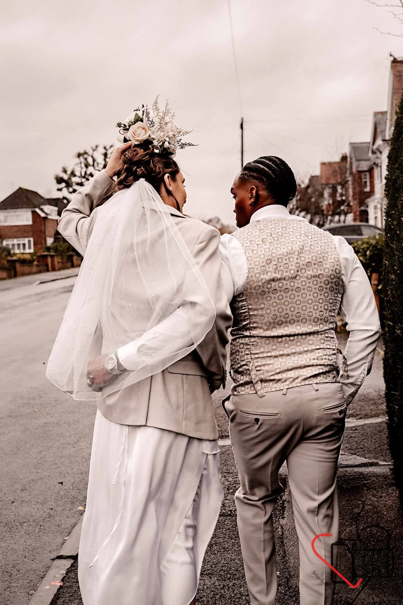A newly married couple stands on a street corner, sharing a moment. The bride is wearing a veil and holding a floral crown above her head, while the groom is dressed in a patterned vest and light-colored pants.