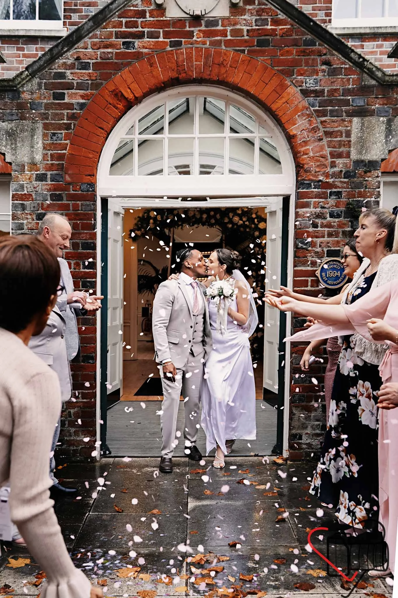 A newlywed couple kissing at their wedding exit as guests throw confetti and petals outside. Portsmouth Register Office, Milldam House.
