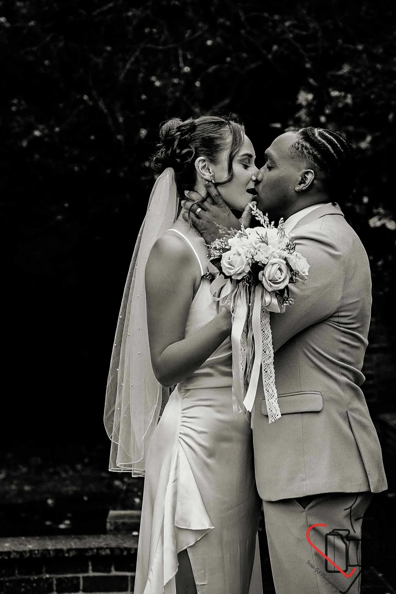 A black and white photograph of a bride and groom kissing, with the bride holding a bouquet of flowers, outdoors with dark foliage in the background.
