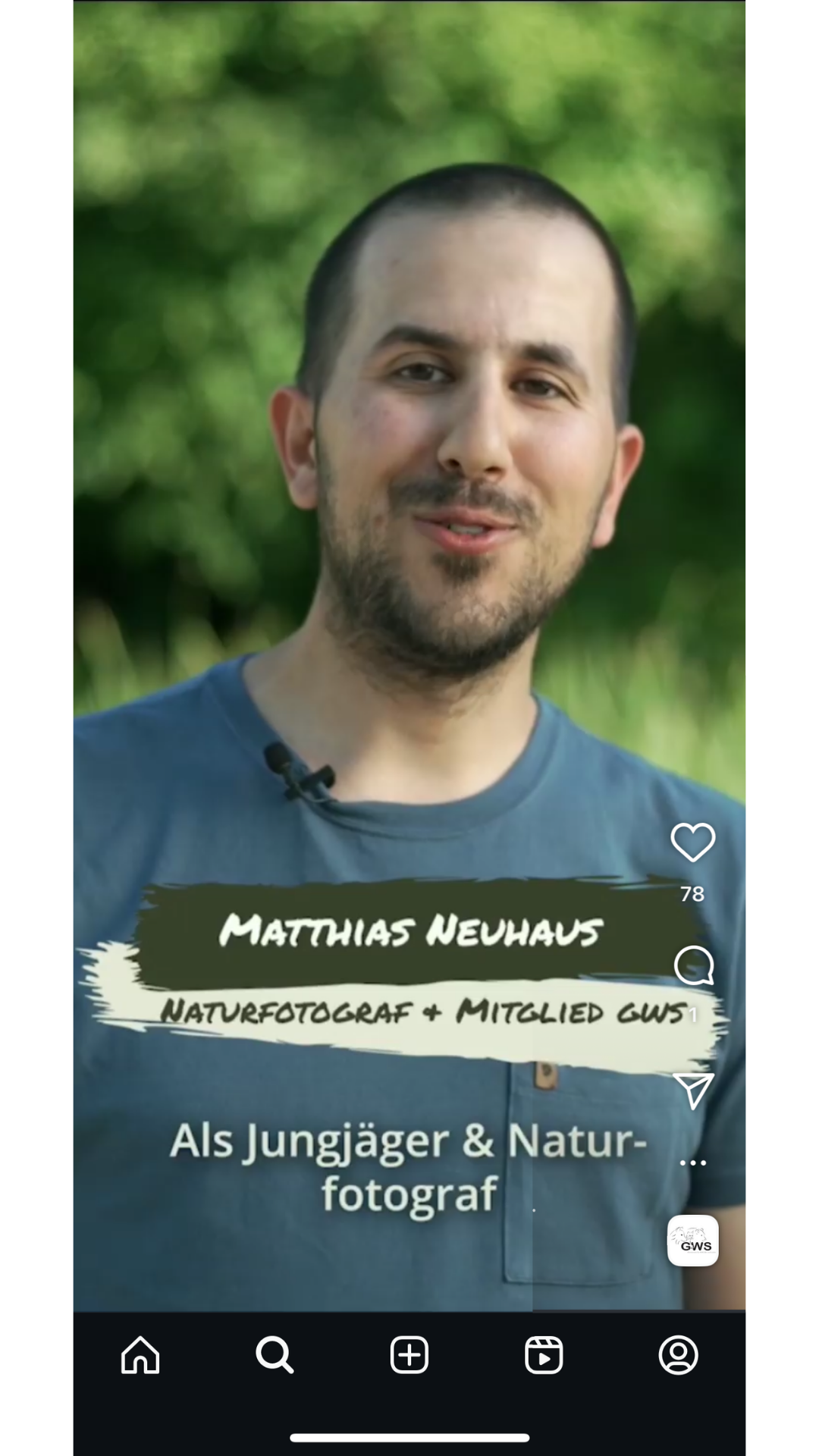 Portrait of a man with short dark hair and a beard, wearing a gray T-shirt with a small pocket, outdoors with green foliage in the background.