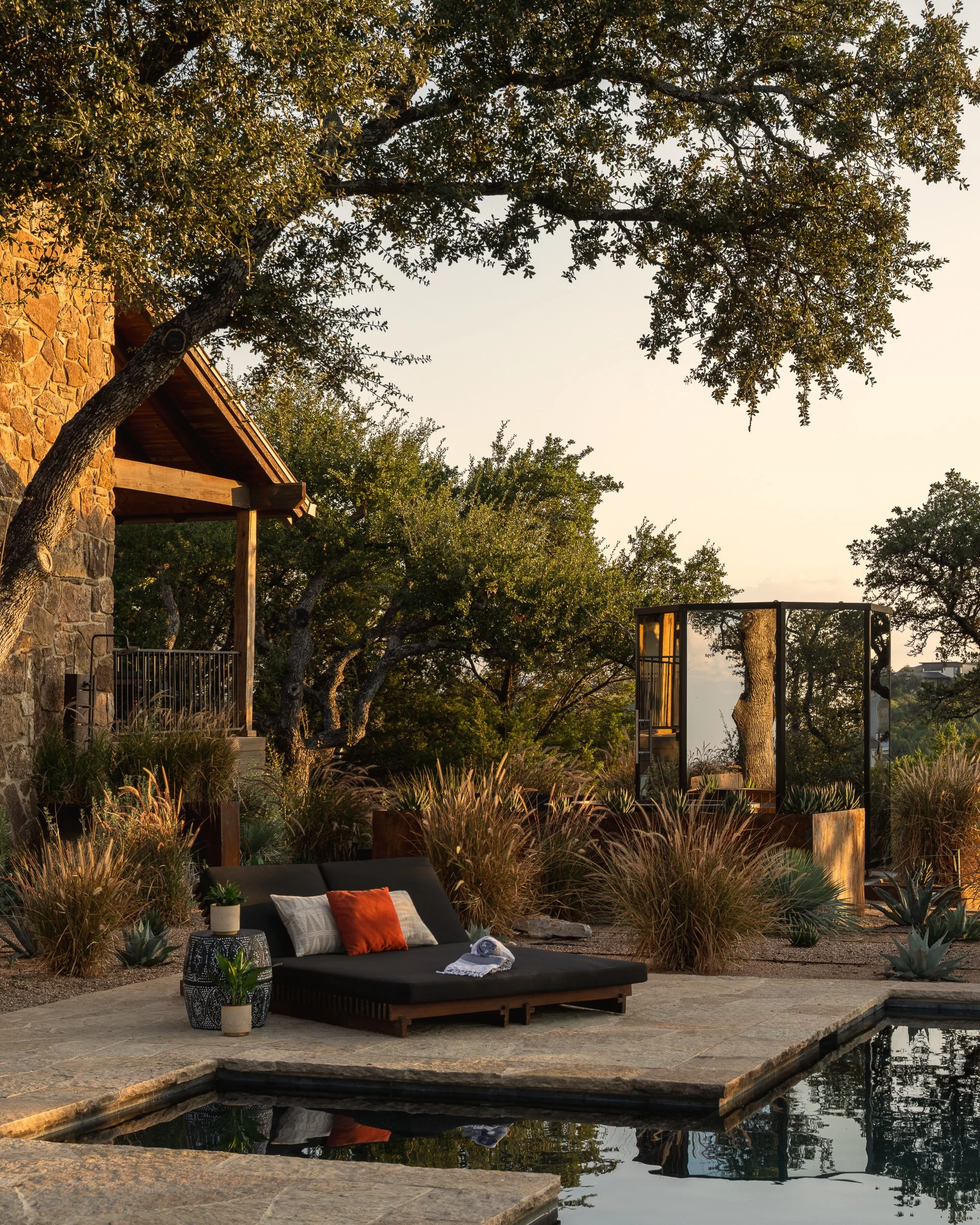 A serene outdoor patio area beside a water feature, with a black lounge chair topped with pillows and a throw, surrounded by potted plants and ornamental grasses. In the background, there are trees, a stone wall, and a structure with a wooden roof, a