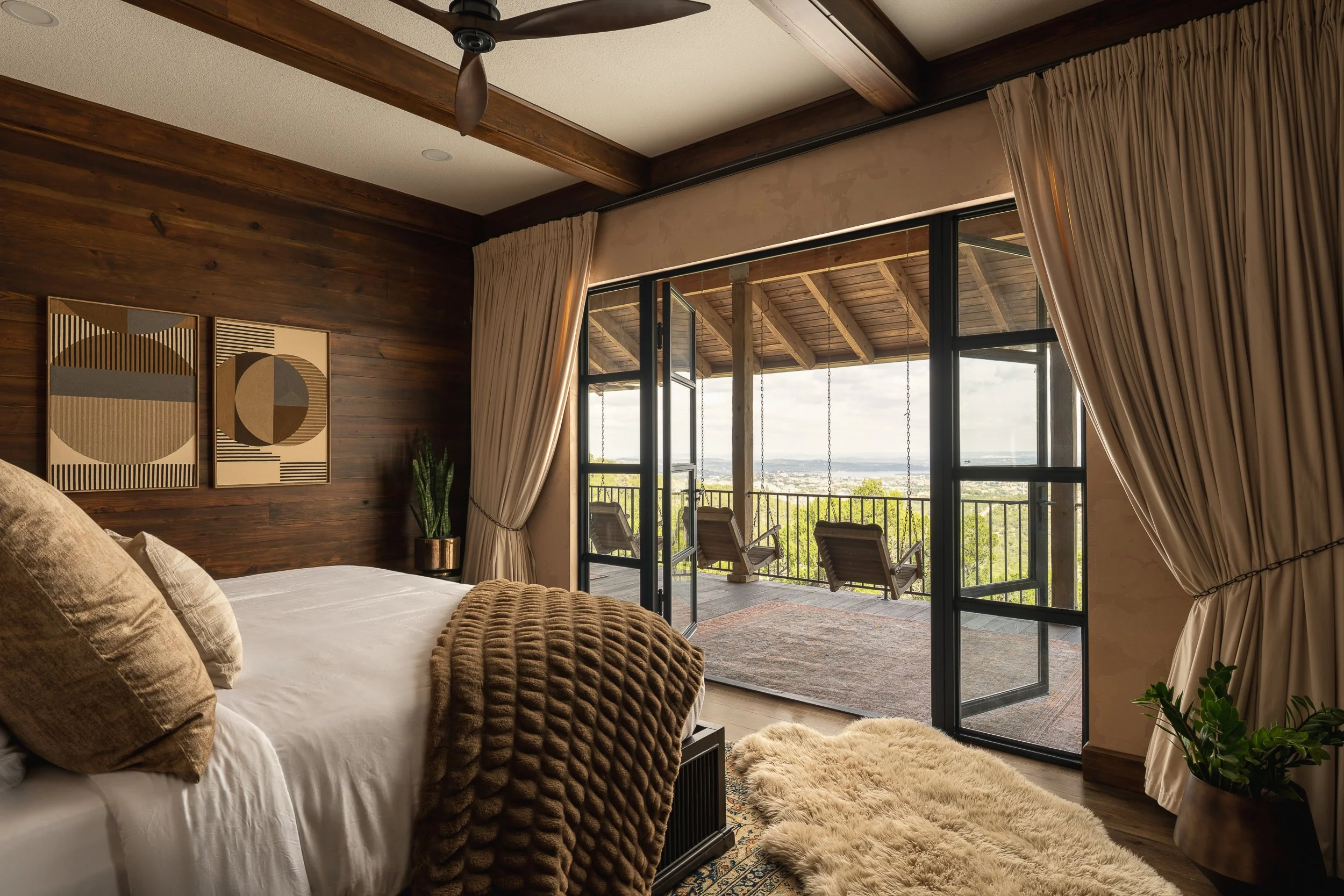 A bedroom with a bed covered in white sheets and beige pillows, a brown knitted blanket, and a textured beige rug. The room has wooden wall paneling, artwork with geometric patterns hanging on the wall, and sliding glass doors that open to a balcony 